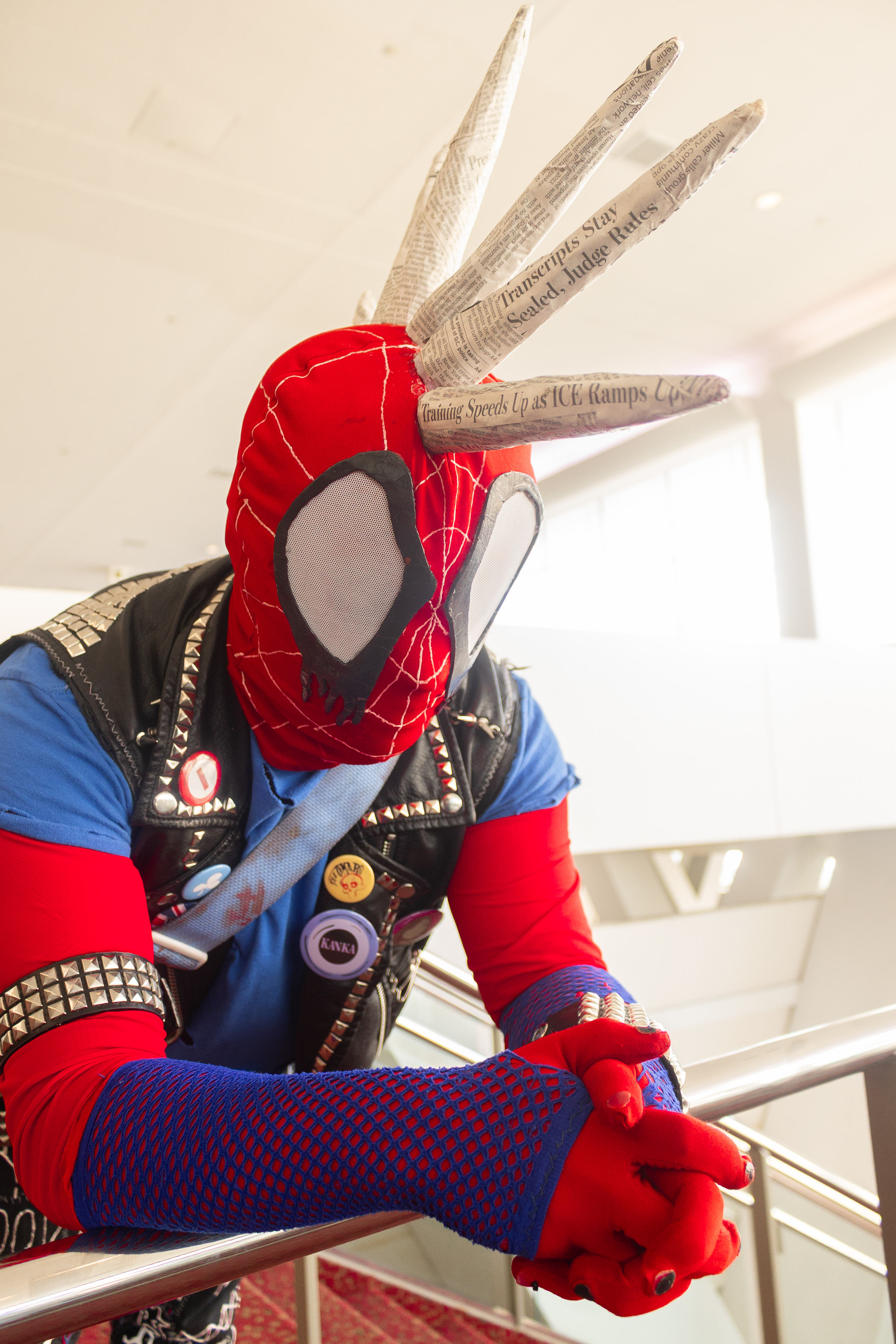 Close-up of Spider-Punk cosplayer with hands clasped on a railing they're leaning on. The mohawk spikes on their mask are covered in newsprint -- two headlines on the front two spikes stand out, one partial "transcripts stay sealed, judge rules" and one complete "training speeds up as ICE ramps up"