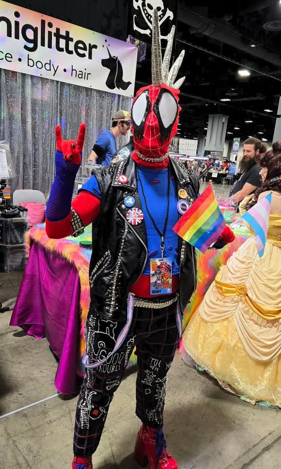Spider-Punk cosplayer holding rainbow and transgender pride flags in one hand and making the "rock on" hand gesture with the other hand