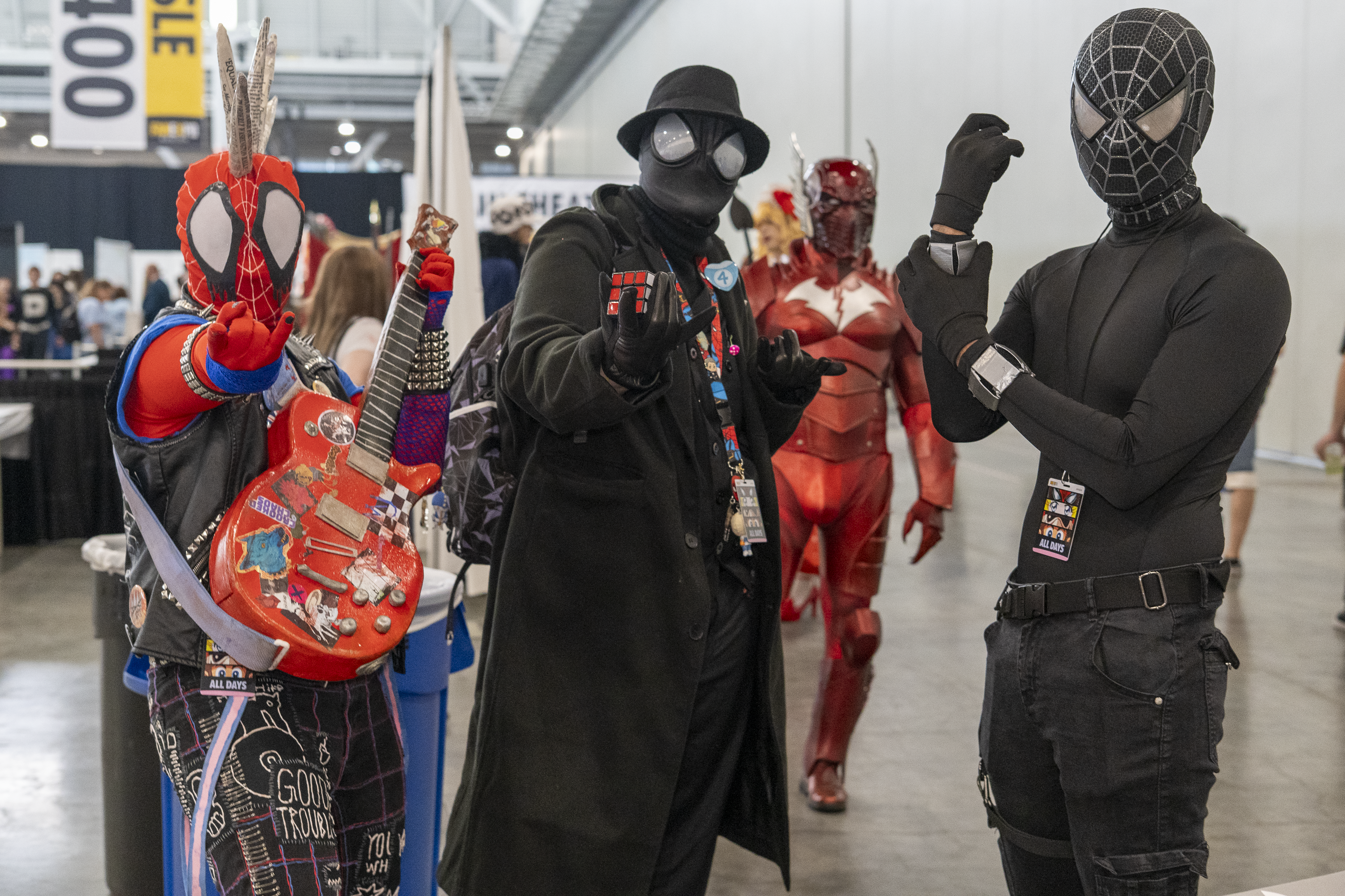 Three Spider-Verse cosplayers in a convention hall. Spider-Punk cosplayer posing with one hand holding their guitar and the other aiming a "web-shooting" hand pose at the camera. Spider-Man Noir cosplayer is holding up a Rubiks Cube. Tactical Spider-Man has fists raised.