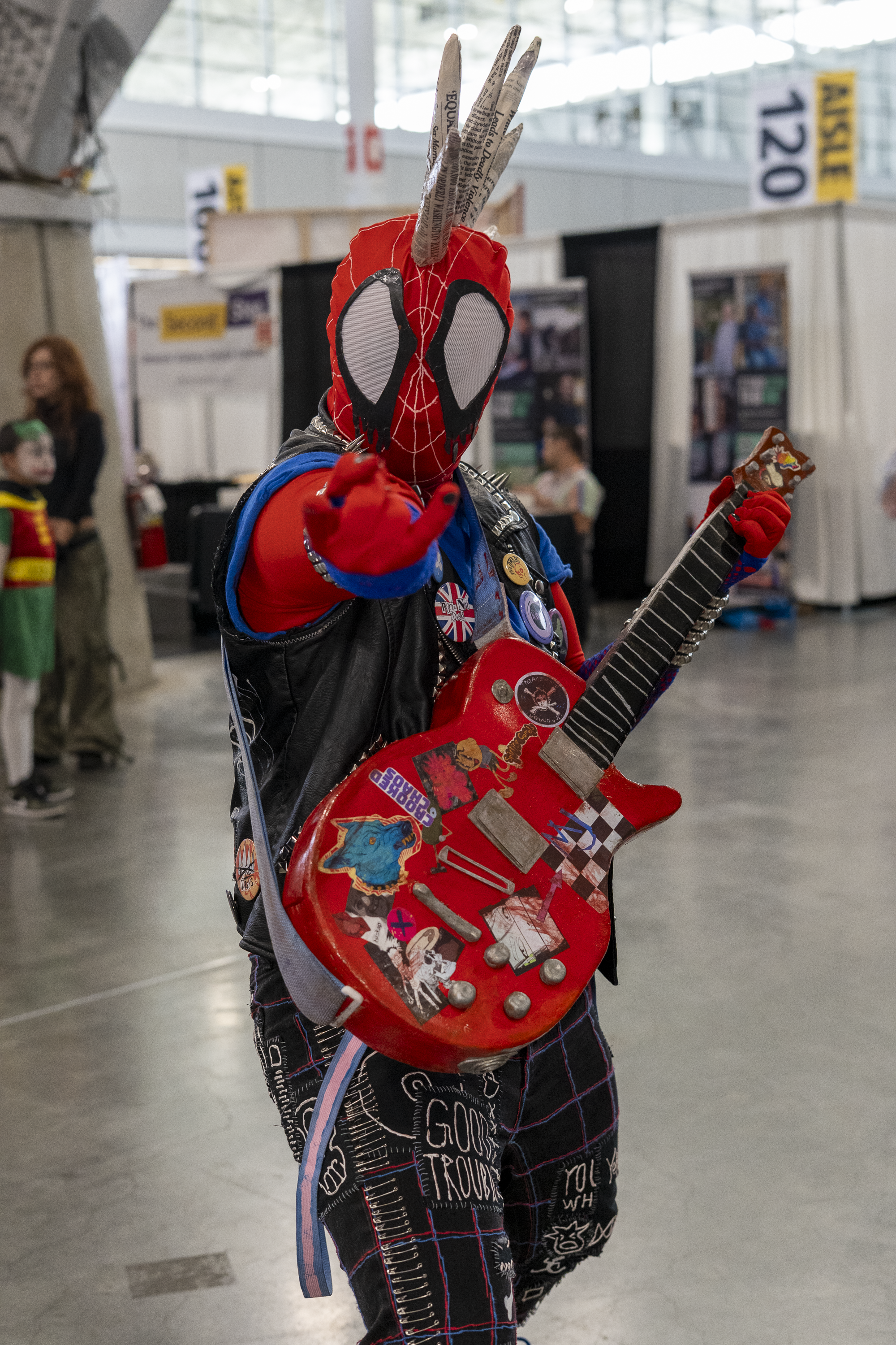 Spider-Punk cosplayer in a convention hall pointing one hand at the camera in a "web-shooting" hand gesture.