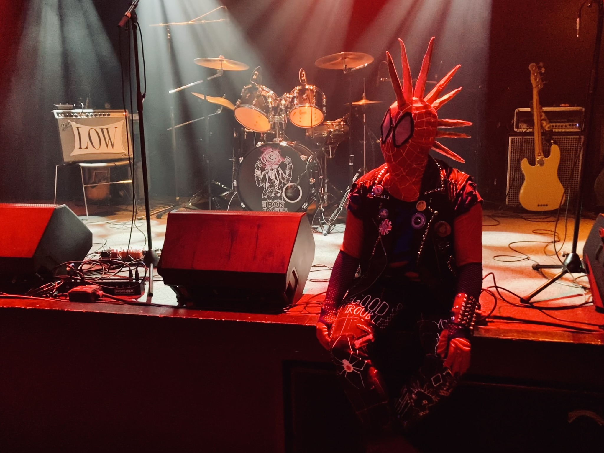 Spider-Punk cosplayer sits on the edge of the stage at a music venue. They are looking towards the top left corner of the frame. The drum set bears the logo of the band The Iron Roses. A spotlight illuminates the "good trouble" patch on their thigh.