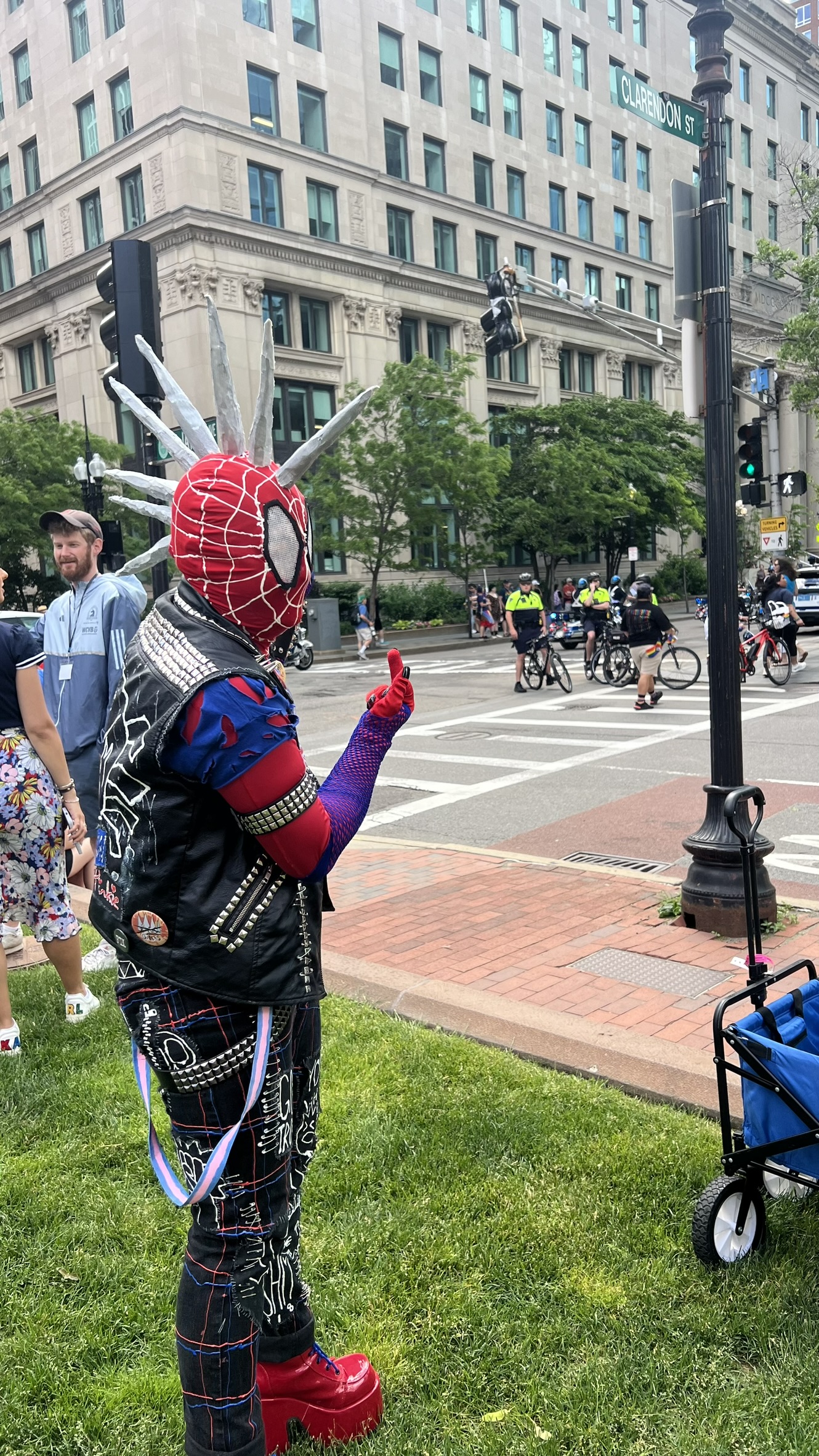 A Spider-Punk cosplayer stands on a grassy patch by a street corner, holding up their middle finger in the direction of a unit of bicycle cops in the intersection.