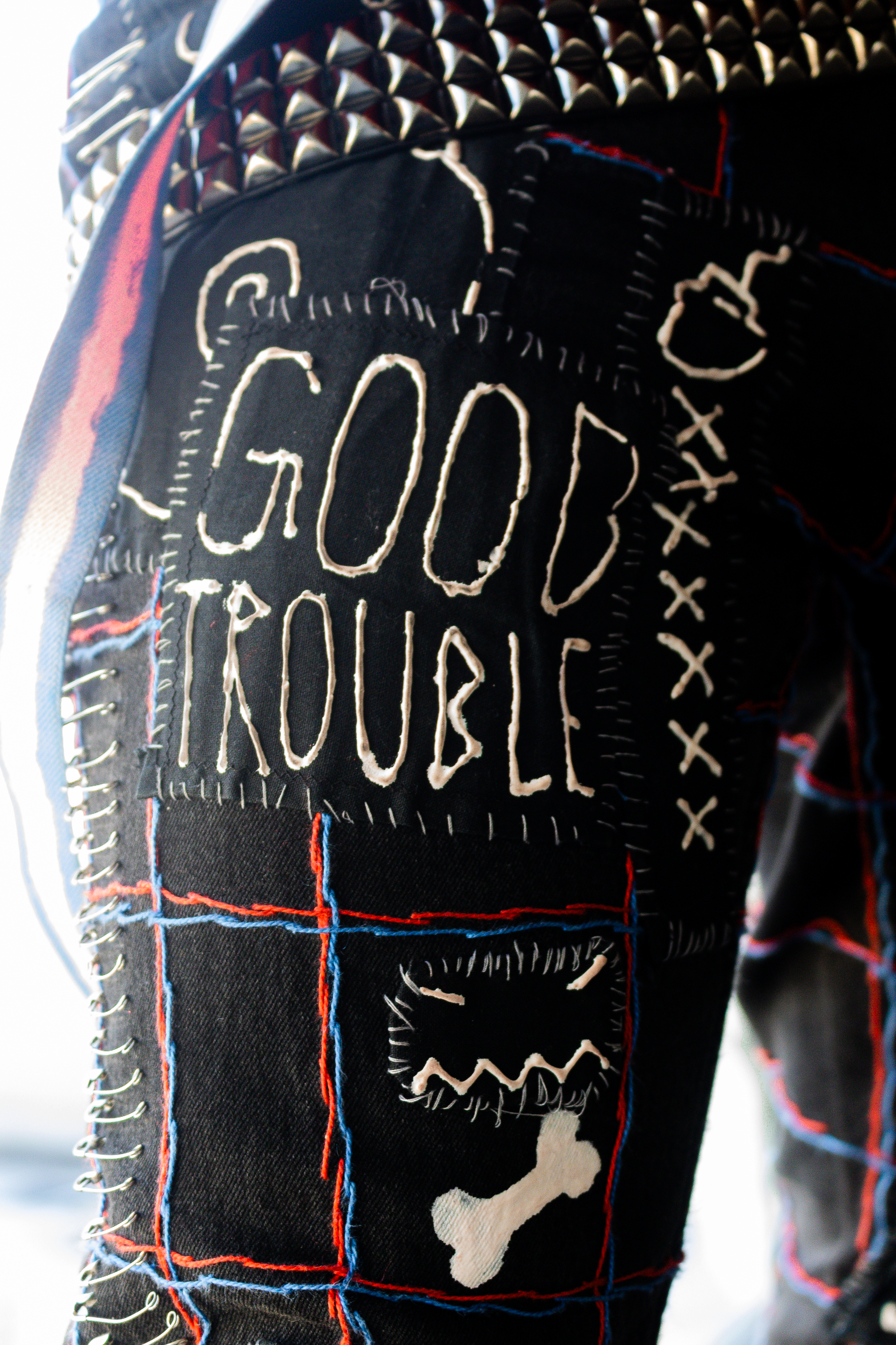Close-up of "Good Trouble" patch on the thigh of the cosplayer's tight-fitting black jeans. The patch is stitched on using perle cotton thread. To the right of the patch is another patch with a bomb and several X's in a vertical line, and below it is a simplistic face made up on=f short diagonal lines for eyes and a zig-zag line for a mouth. The jeans are also embroidered with a grid pattern with blue and red lines parallel to one another going both vertically and horizontally. Visible on the outer hip is the cosplayer's pink-and-blue suspenders, which are not worn properly but hanging from their belt.