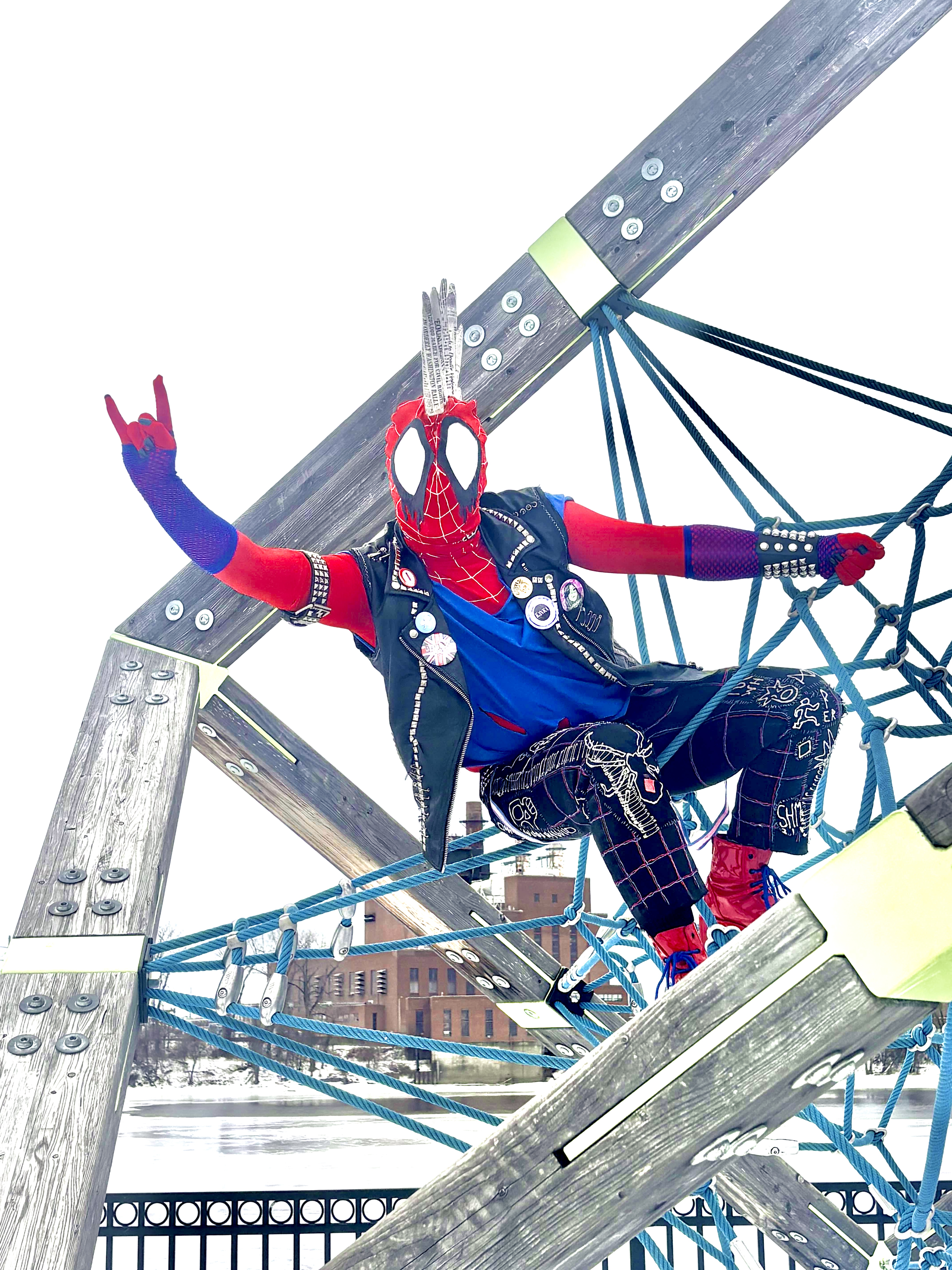 A Spider-Punk cosplayer has climbed up a playground structure best described as a rope web. They are sitting on the web and holding on to it with one hand, while leaning far out from it and holding their free hand out in a "rock on" hand gesture
