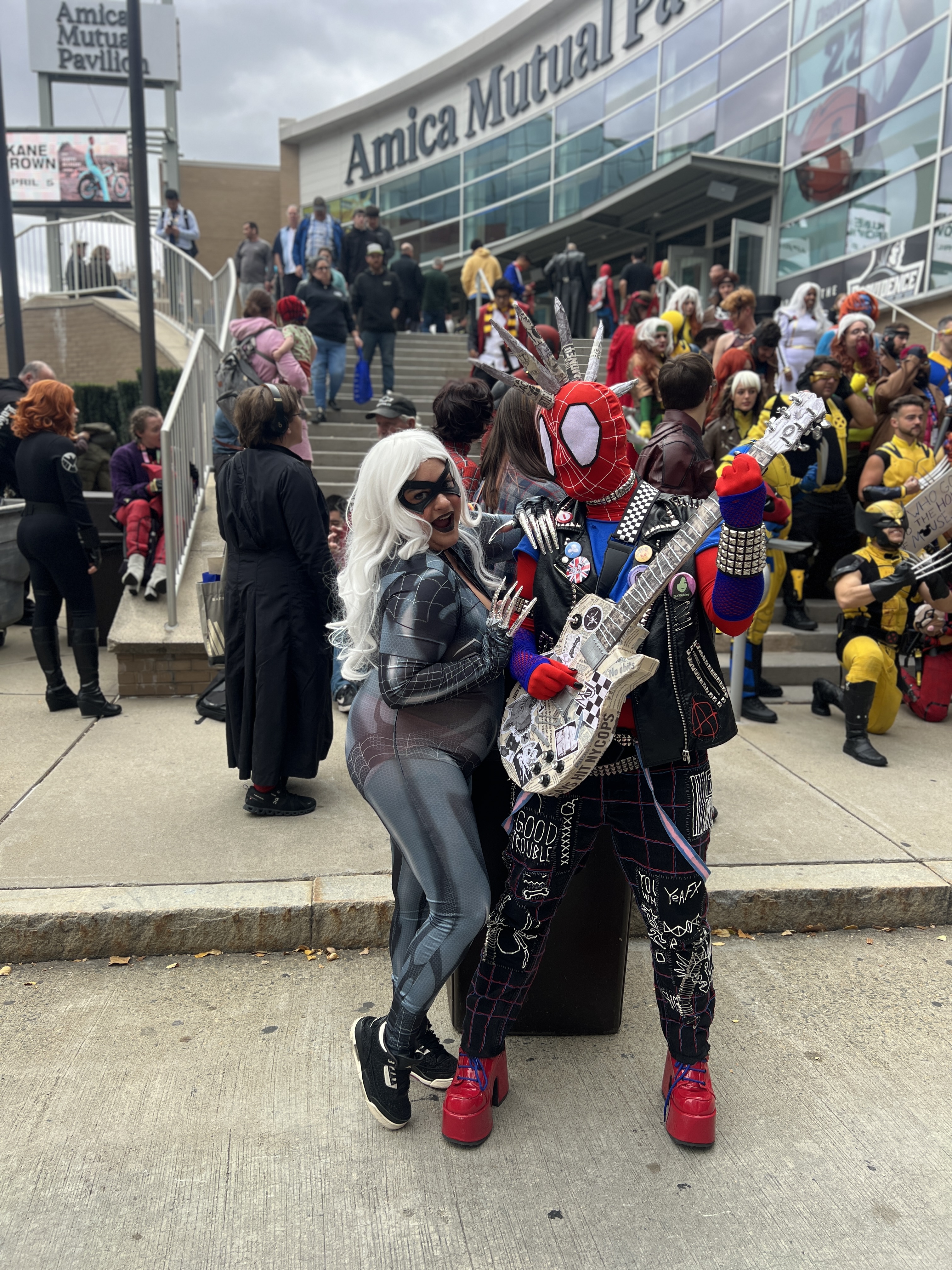 Spider-Punk and Black Cat cosplayers posing together outside the Amica Mutual Pavilion. In the background, X-Men cosplayers are gathered on the stairs.