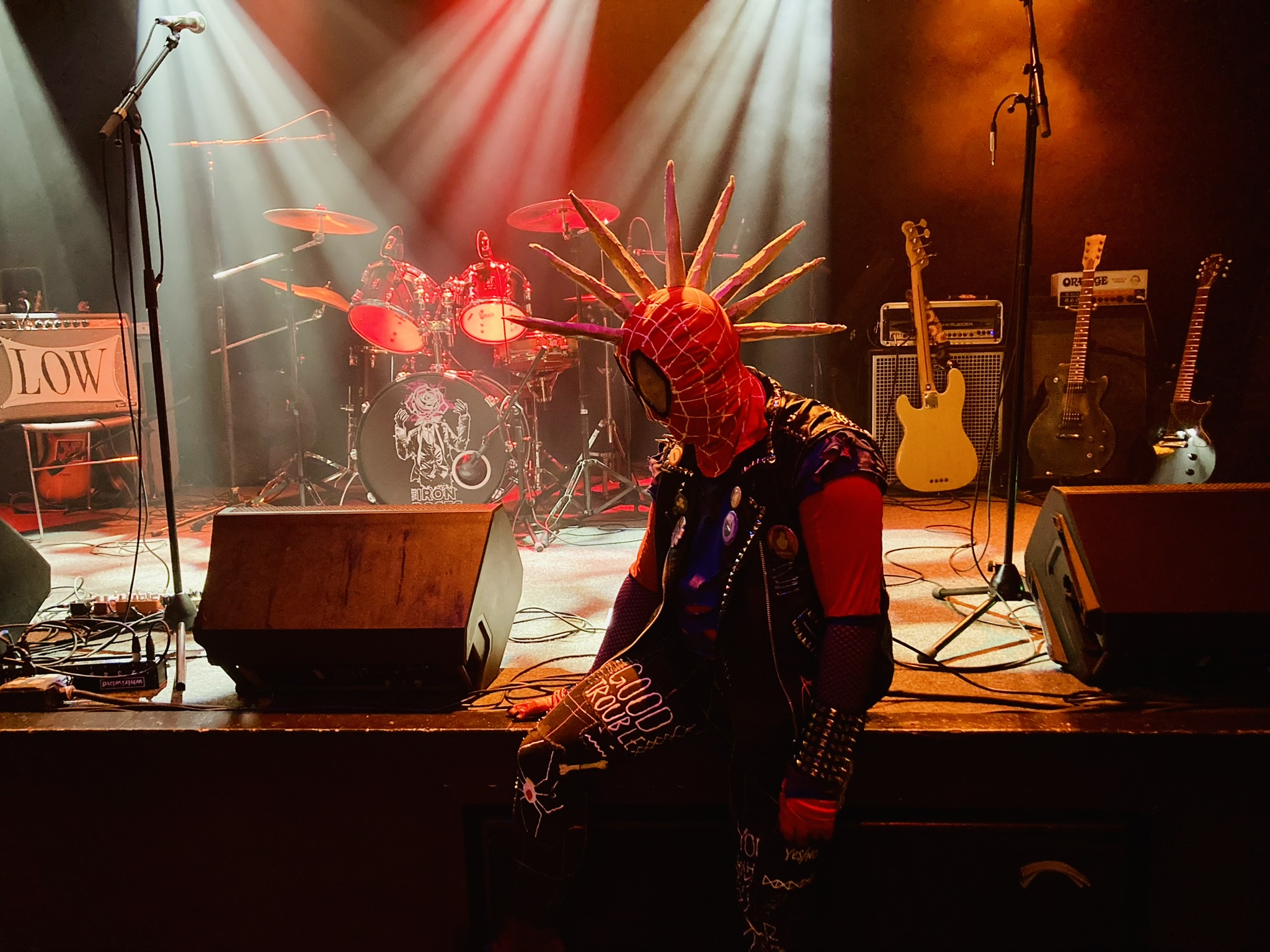 Spider-Punk cosplayer sits on the edge of the stage at a music venue. They are looking towards the bottom left corner of the stage. The drum set bears the logo of the band The Iron Roses. A spotlight illuminates the "good trouble" patch on their thigh.