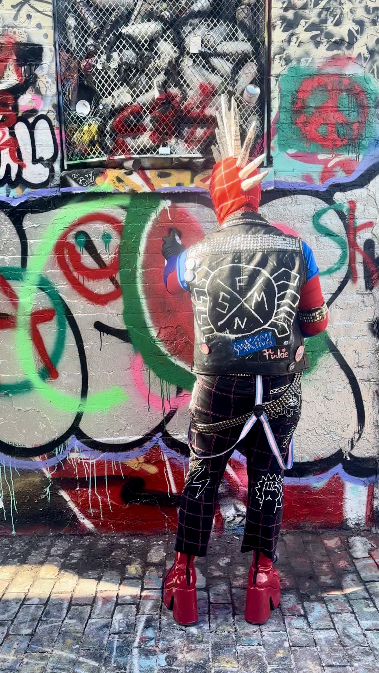 A Spider-Punk cosplayer faces away from the camera in front of a heavily graffitied wall, spray-painting a large watermelon on it.