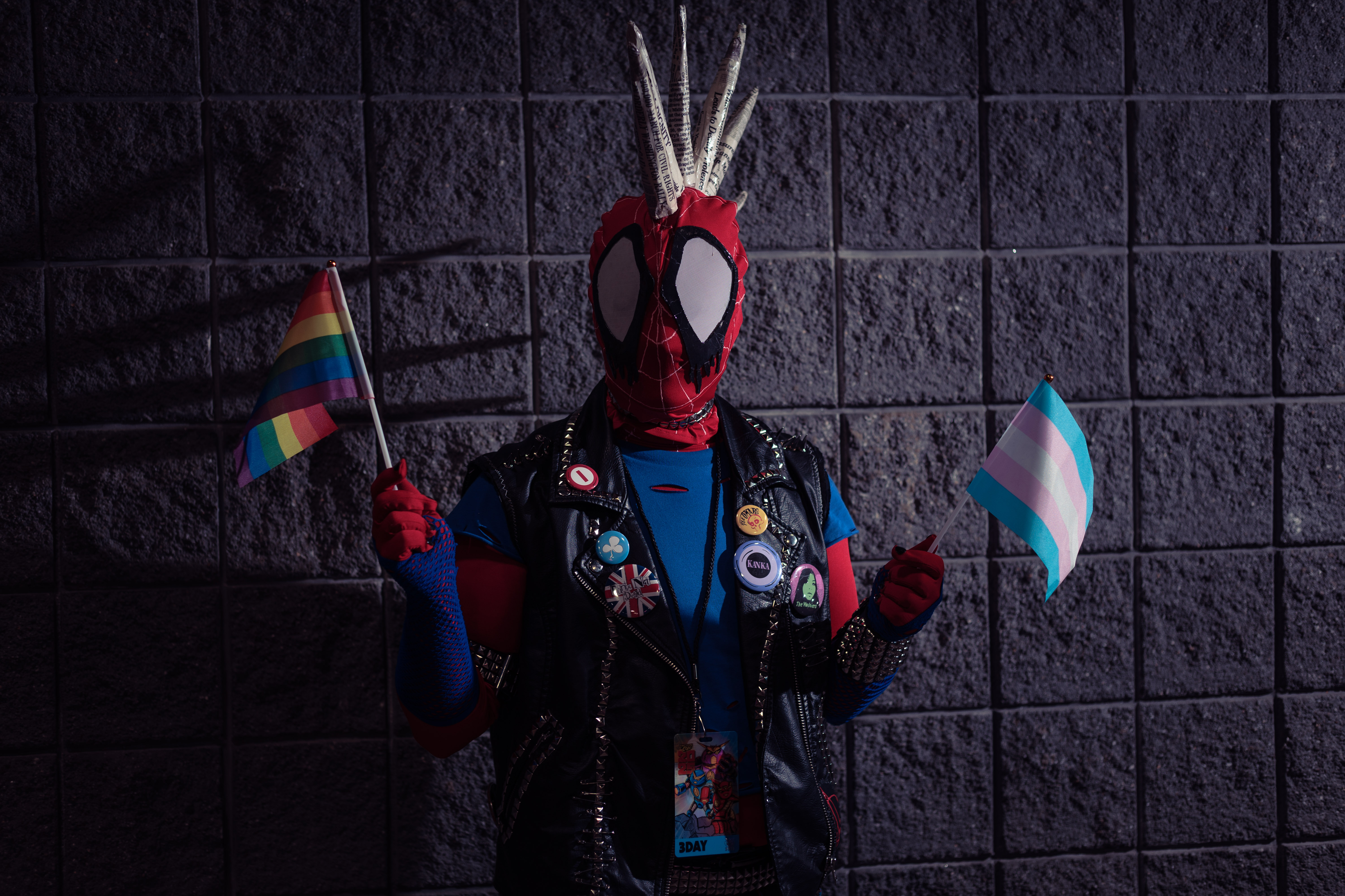 A Spider-Punk cosplayer with newsprint-covered mask mohawk spikes stands in front of a wall holding small rainbow and transgender pride flags, one in each hand