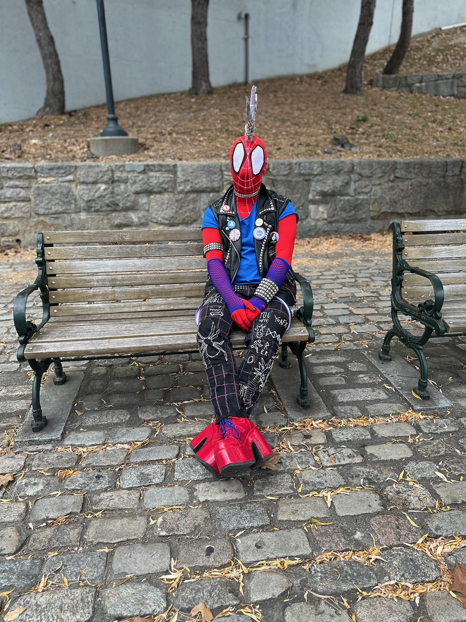 A Spider-Punk cosplayer wearing red platform heeled boots sits on a bench, legs crossed and hands clasped in their lap.