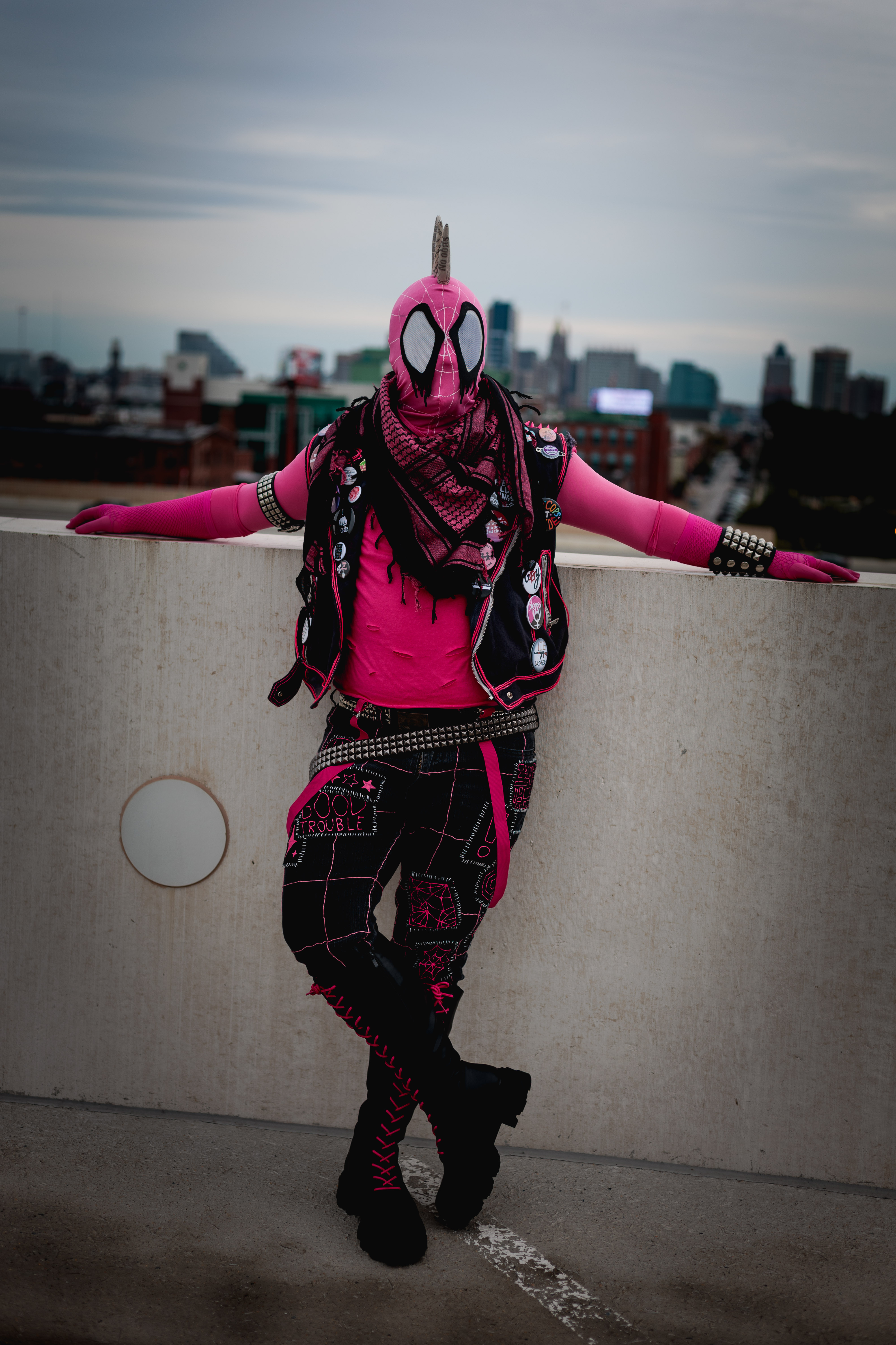 Spider-Punk (pink palette) cosplayer wearing a pink keffiyeh, leaning against the wall on the top floor of a garage. The sky is cloudy