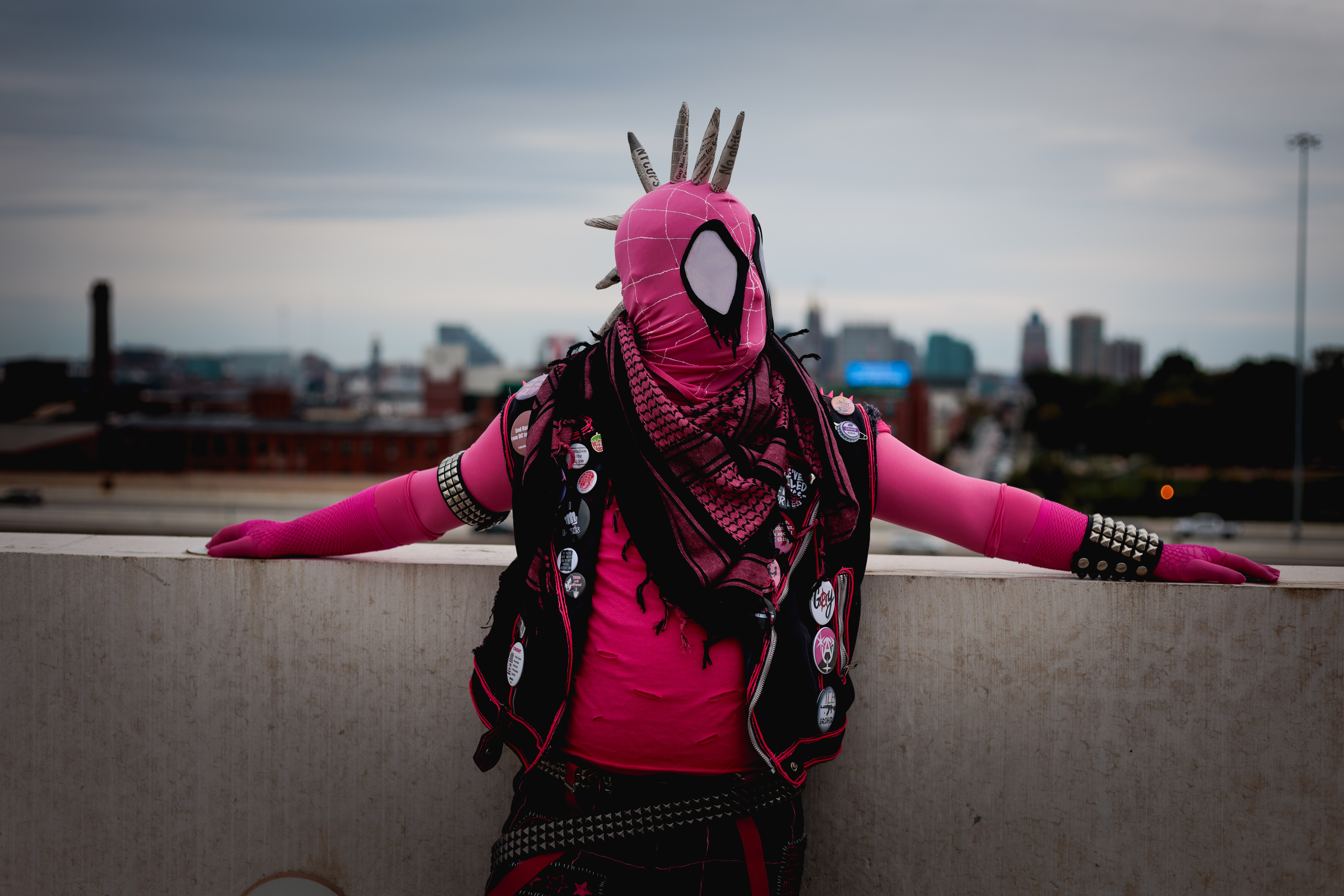 Spider-Punk (pink palette) cosplayer wearing a pink keffiyeh, leaning against the wall on the top floor of a garage. The sky is cloudy