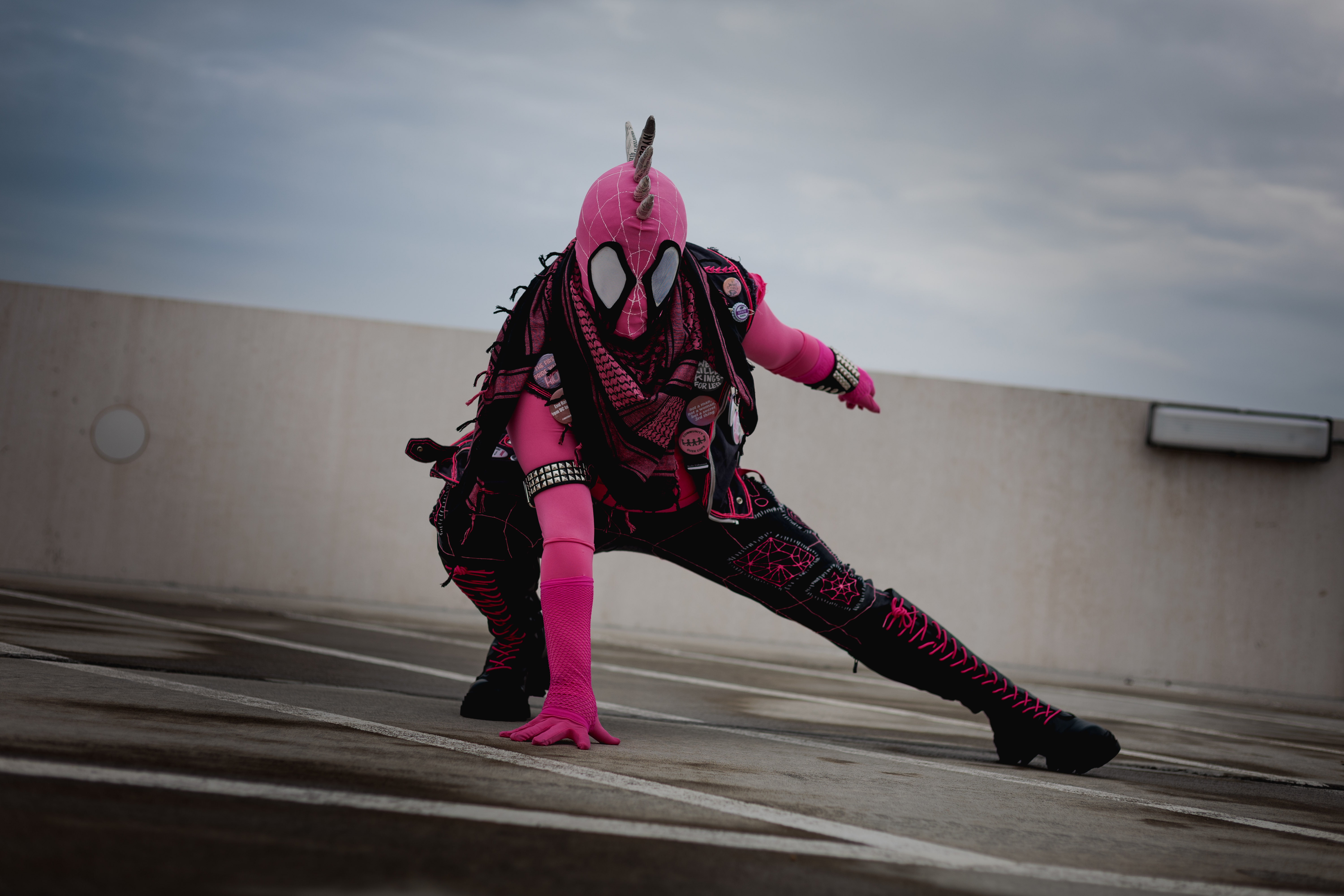 Spider-Punk (pink palette) cosplayer wearing a pink keffiyeh, in the classic Spider-Man crouch pose. The sky is cloudy