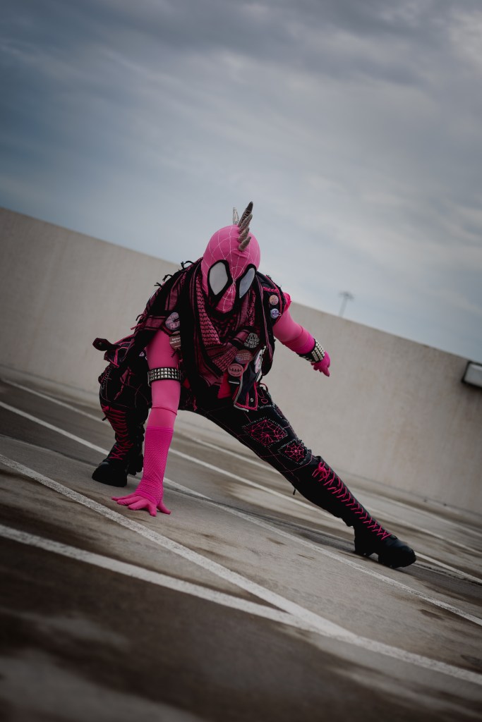 Spider-Punk (pink palette) cosplayer wearing a pink keffiyeh, in the classic Spider-Man crouch pose. The sky is cloudy