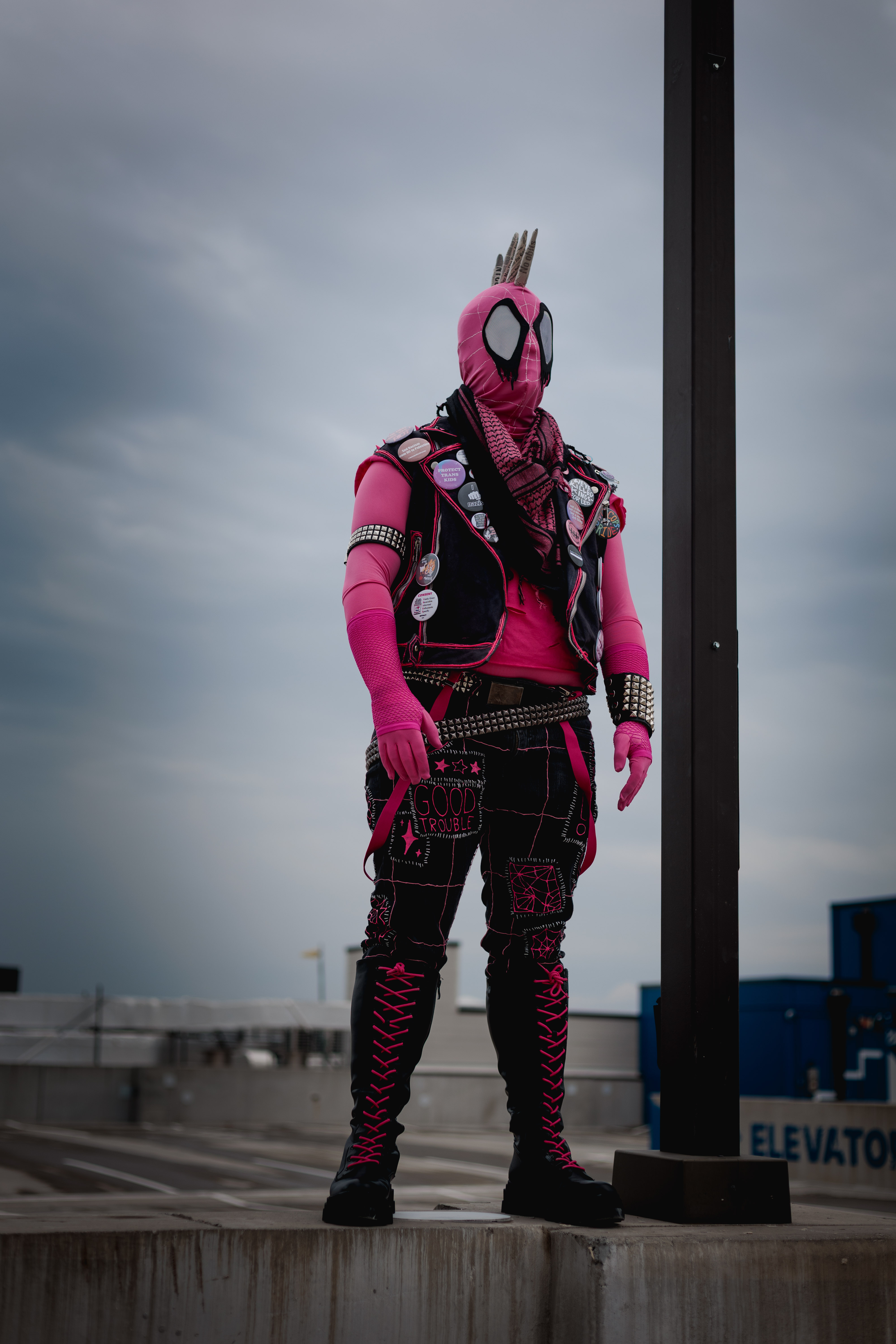 Spider-Punk (pink palette) cosplayer wearing a pink keffiyeh, standing underneath a lamp post on the top floor of a garage. The sky is cloudy