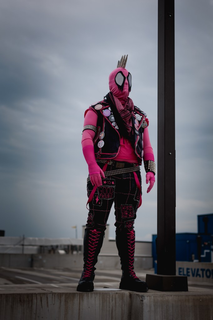 Spider-Punk (pink palette) cosplayer wearing a pink keffiyeh, standing underneath a lamp post on the top floor of a garage. The sky is cloudy