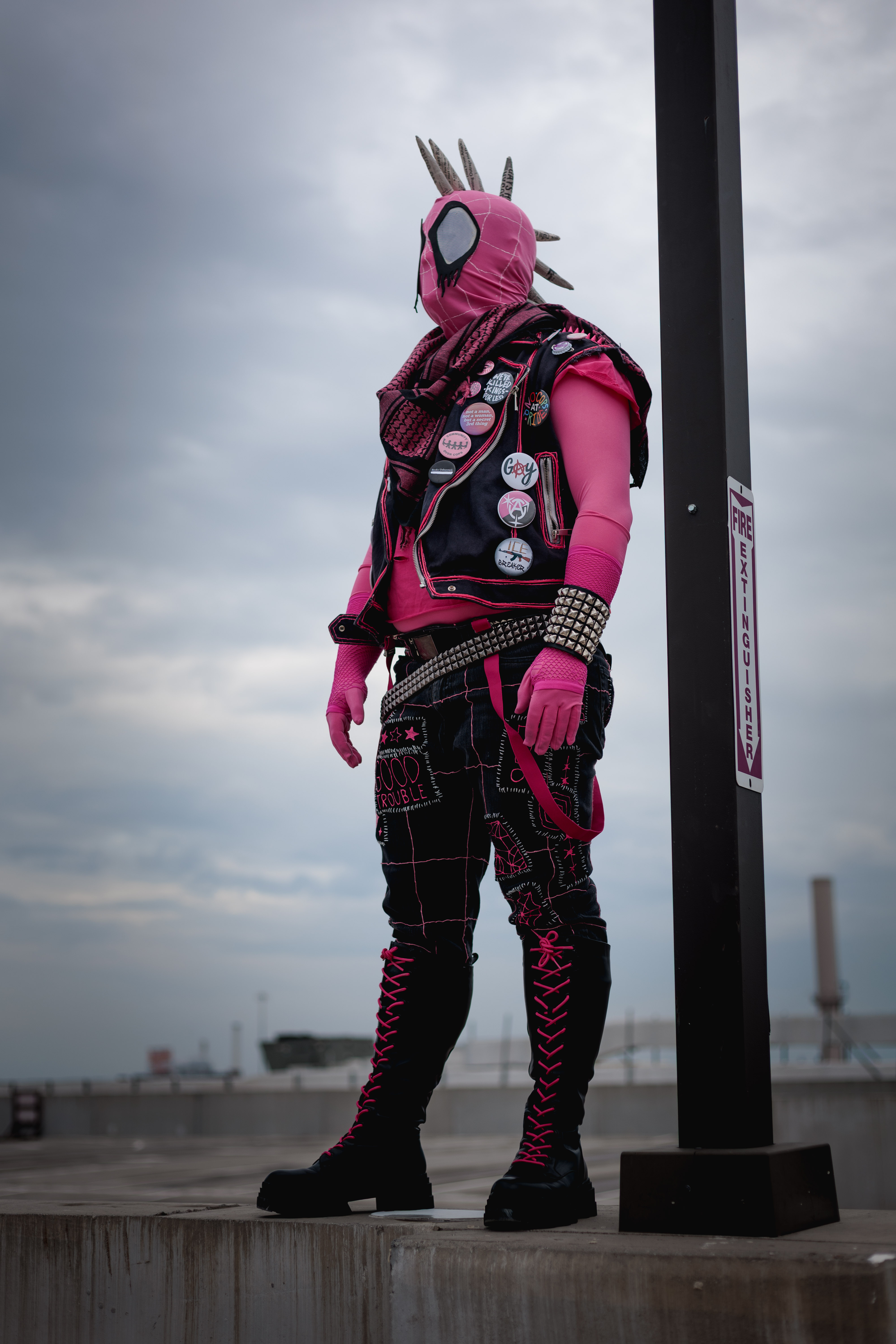 Spider-Punk (pink palette) cosplayer wearing a pink keffiyeh, standing underneath a lamp post on the top floor of a garage. The sky is cloudy