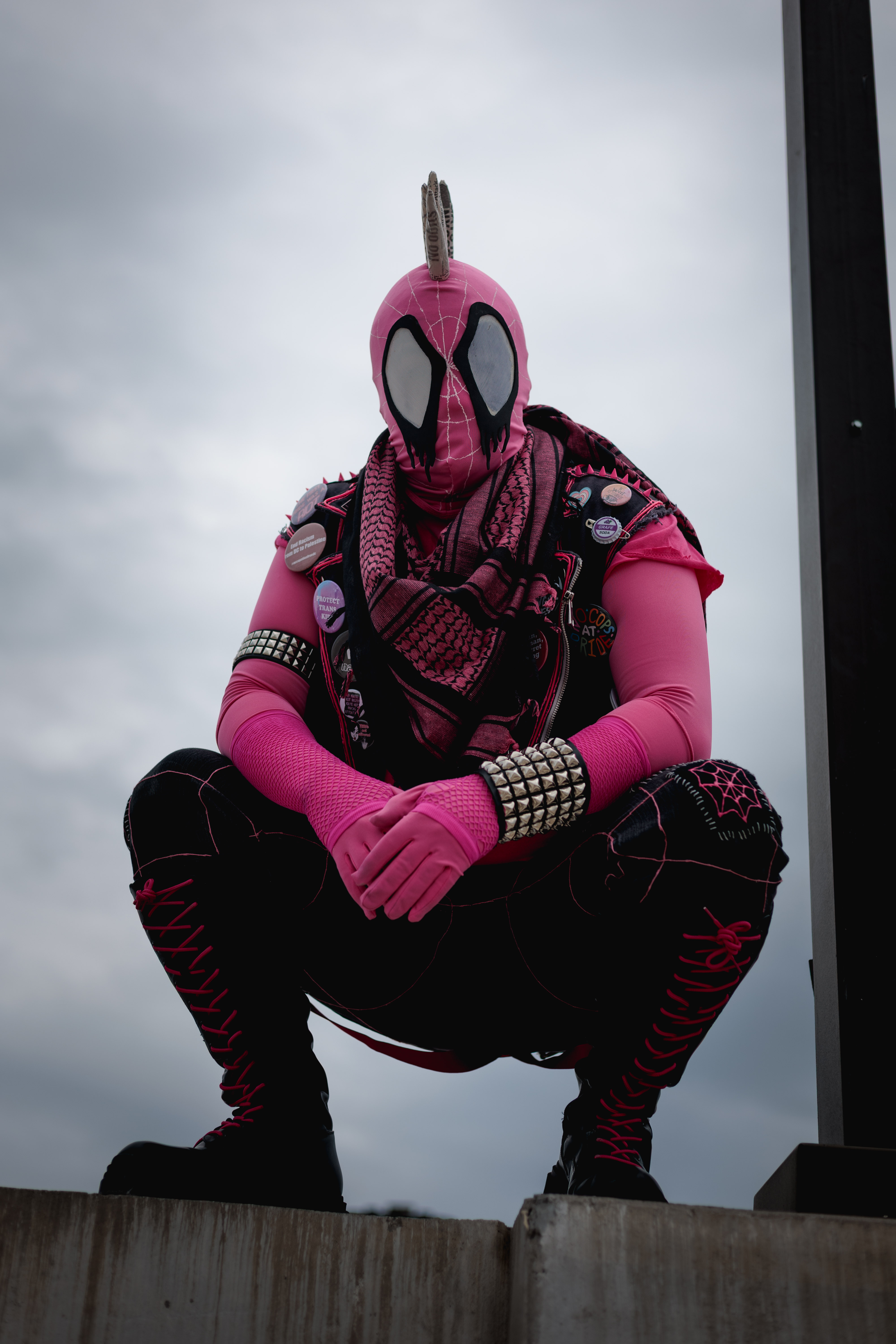 Spider-Punk (pink palette) cosplayer wearing a pink keffiyeh, squatting underneath a lamp post on the top floor of a garage. The sky is cloudy