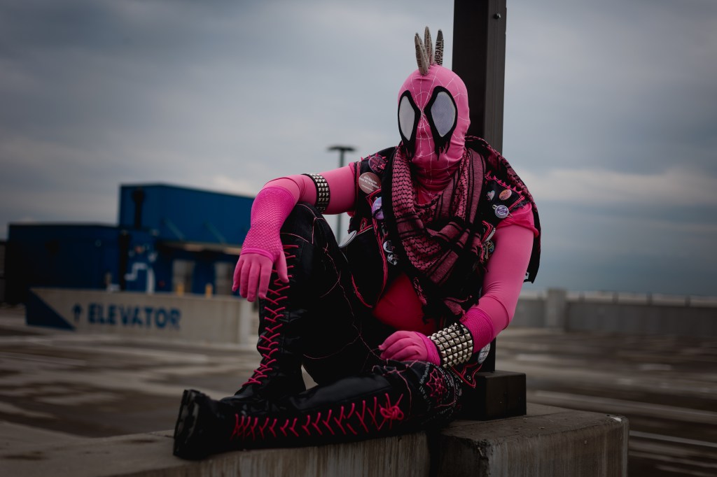 Spider-Punk (pink palette) cosplayer wearing a pink keffiyeh, sitting underneath a lamp post on the top floor of a garage. The sky is cloudy