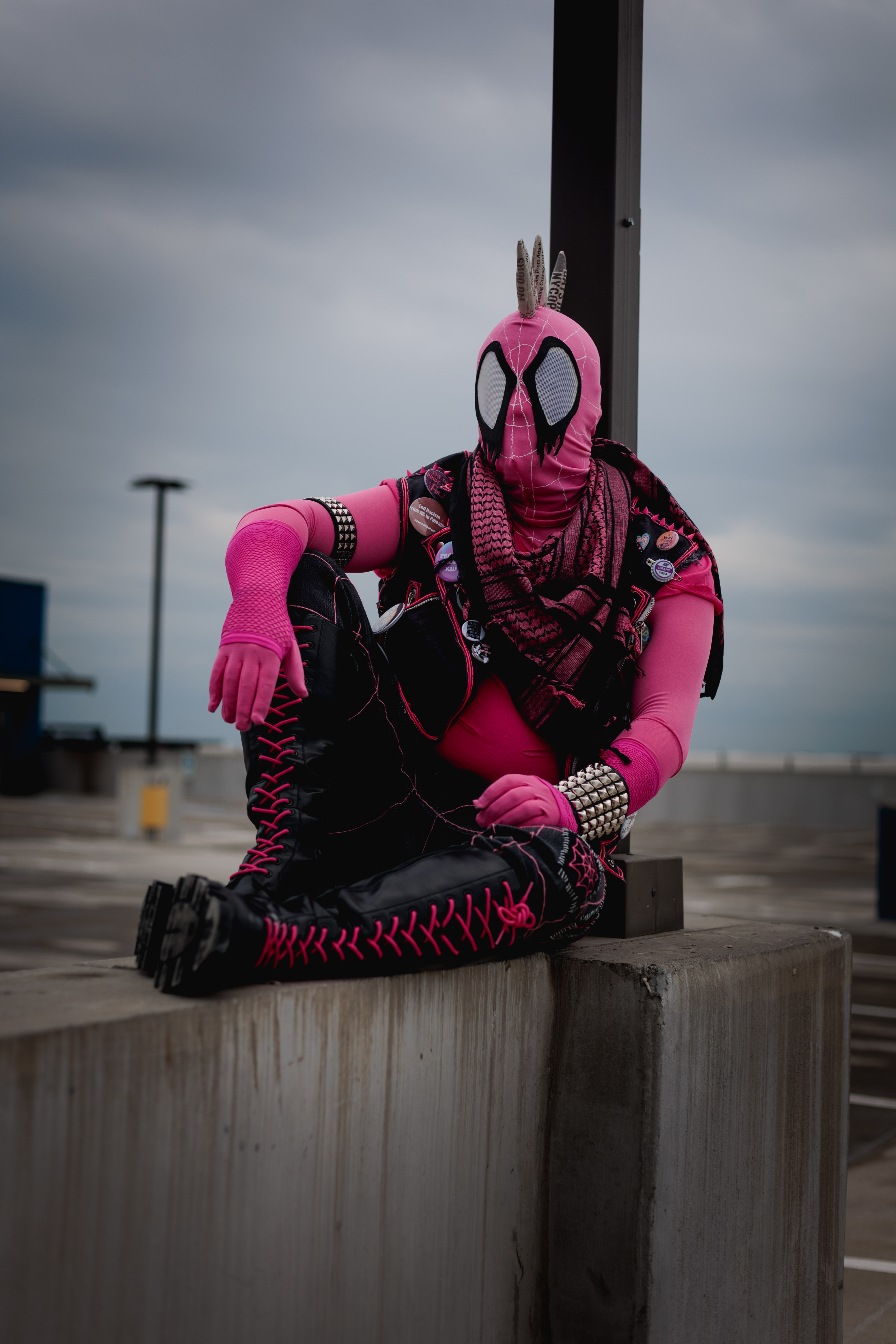 Spider-Punk (pink palette) cosplayer wearing a pink keffiyeh, sitting underneath a lamp post on the top floor of a garage. The sky is cloudy