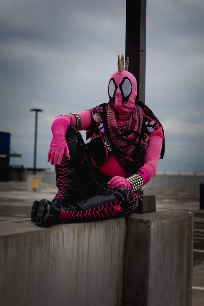 Spider-Punk (pink palette) cosplayer wearing a pink keffiyeh, sitting underneath a lamp post on the top floor of a garage. The sky is cloudy
