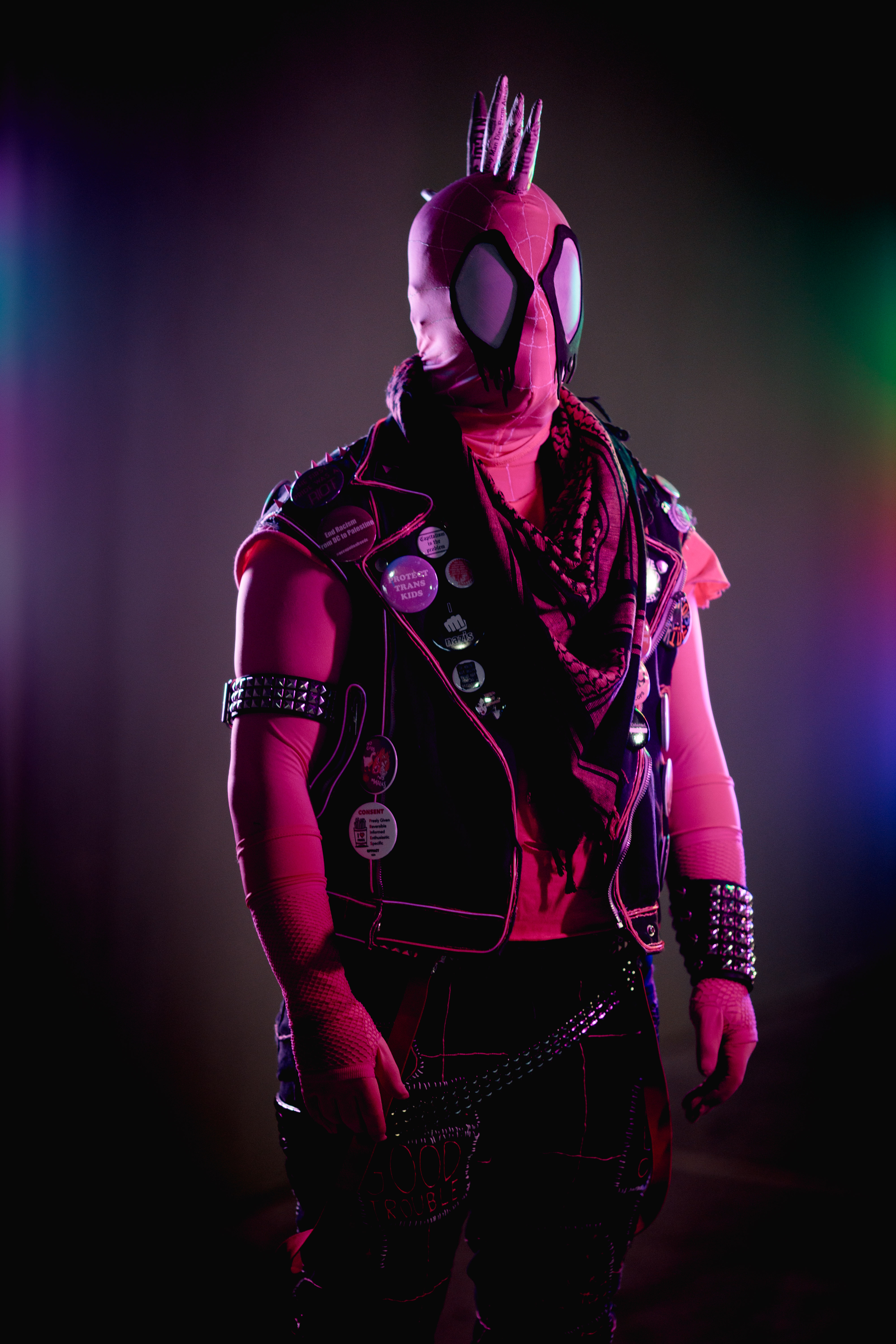 Spider-punk (pink palette) cosplayer wearing a pink keffiyeh. Lighting on the left side of the photo is transgender flag colors. Lighting on the right side is rainbow colors.