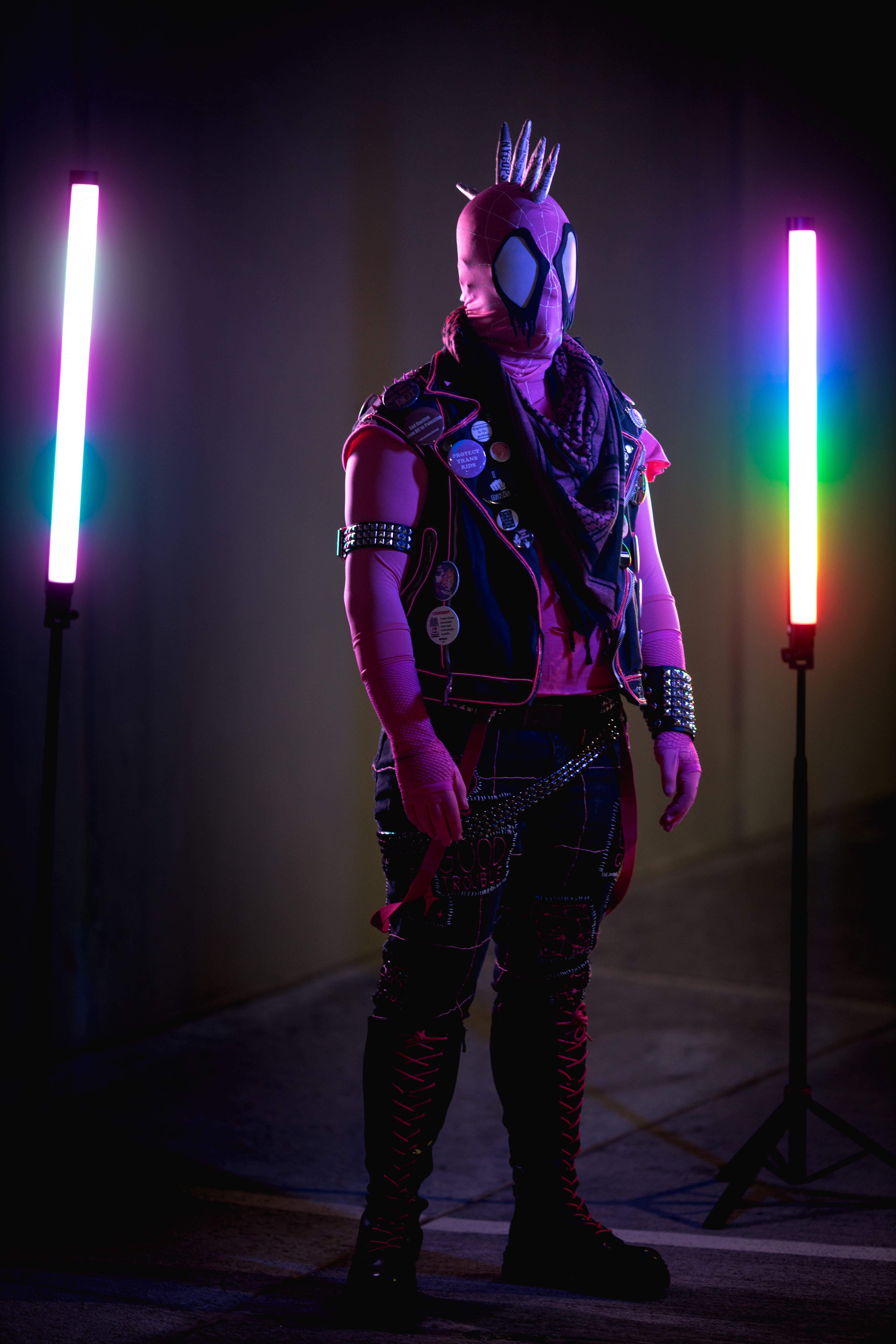 Spider-punk (pink palette) cosplayer wearing a pink keffiyeh, illuminated by light tubes on either side. The left tube displays transgender flag colors, and the right tube displays rainbow colors.