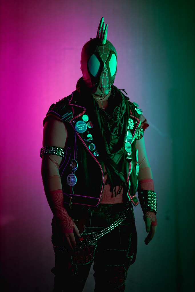 Spider-punk (pink palette) cosplayer wearing a pink keffiyeh illuminated by colorful light