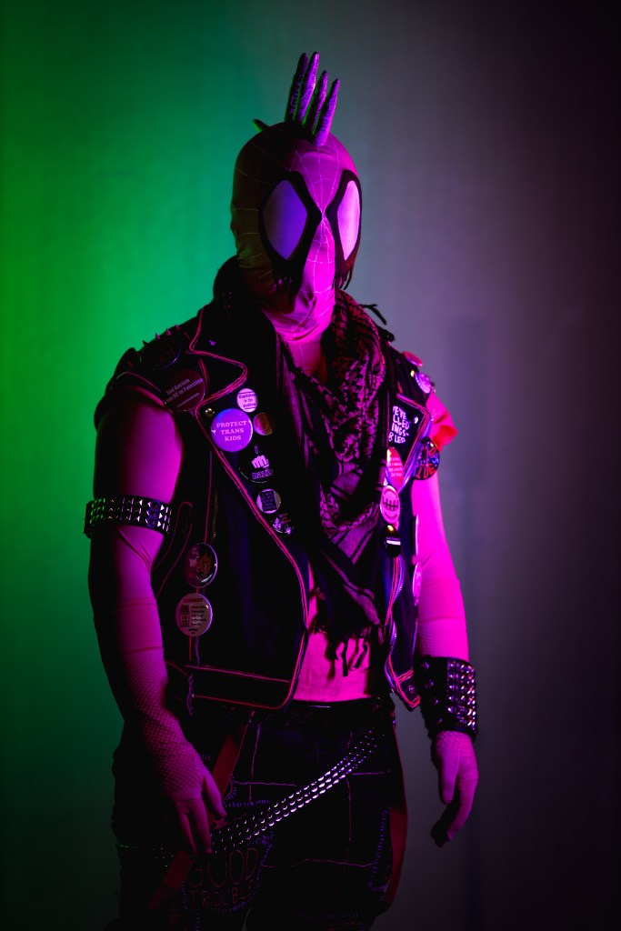 Spider-punk (pink palette) cosplayer wearing a pink keffiyeh, illuminated by colorful light