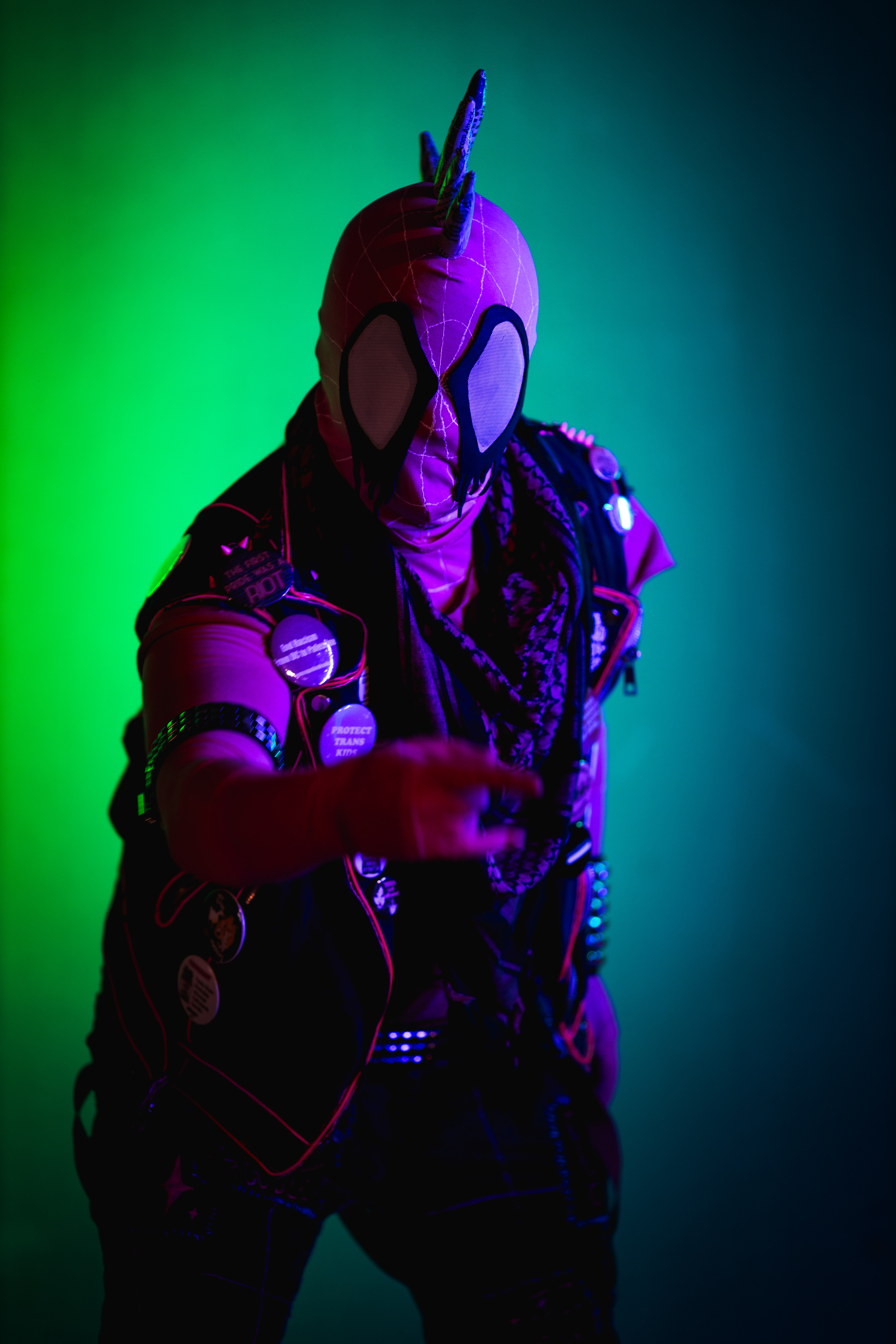 Spider-punk (pink palette) cosplayer wearing a pink keffiyeh illuminated by colorful light, pointing a "web-shooting" hand gesture at the camera
