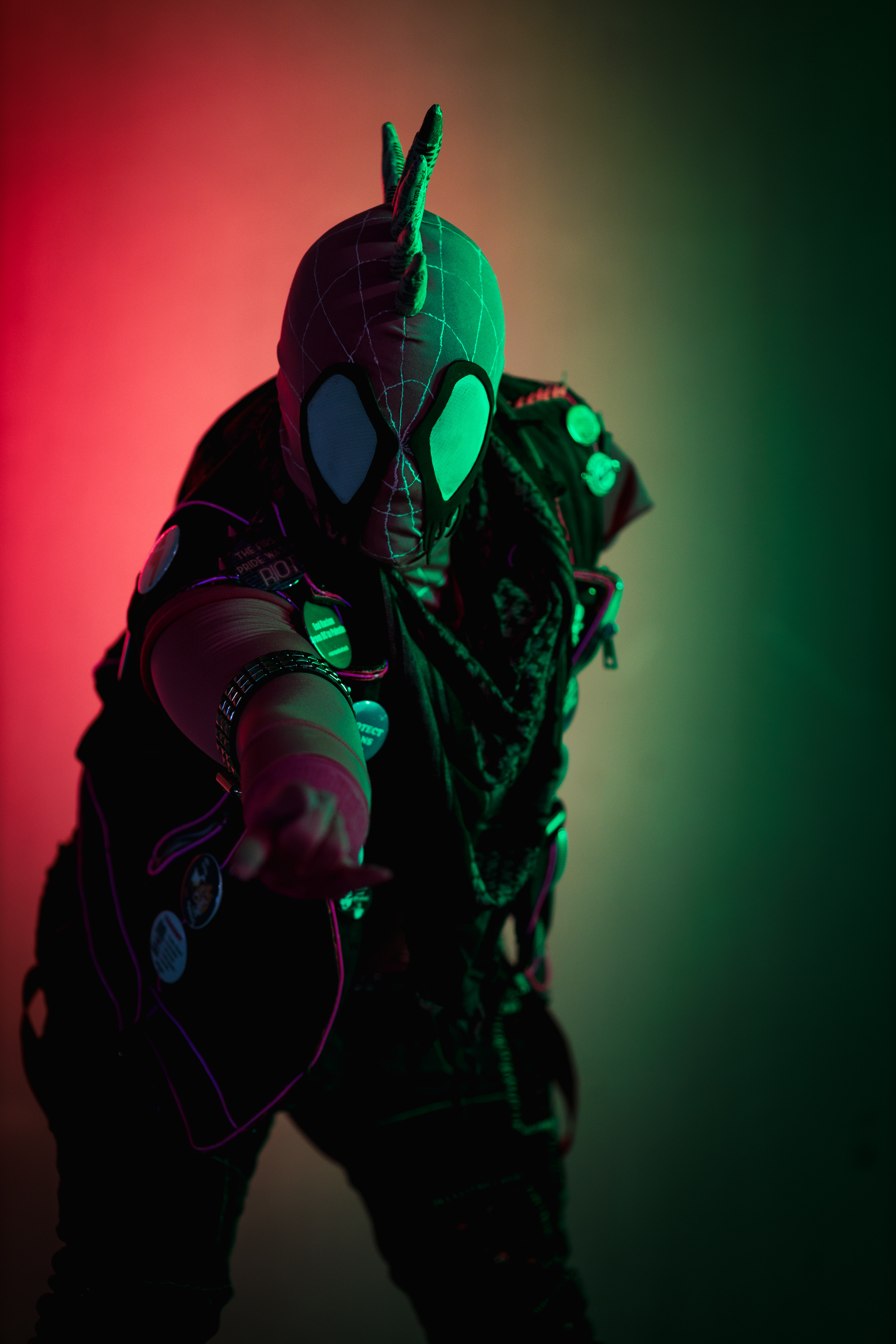 Spider-punk (pink palette) cosplayer wearing a pink keffiyeh illuminated by colorful light, pointing a "web-shooting" hand gesture at the camera