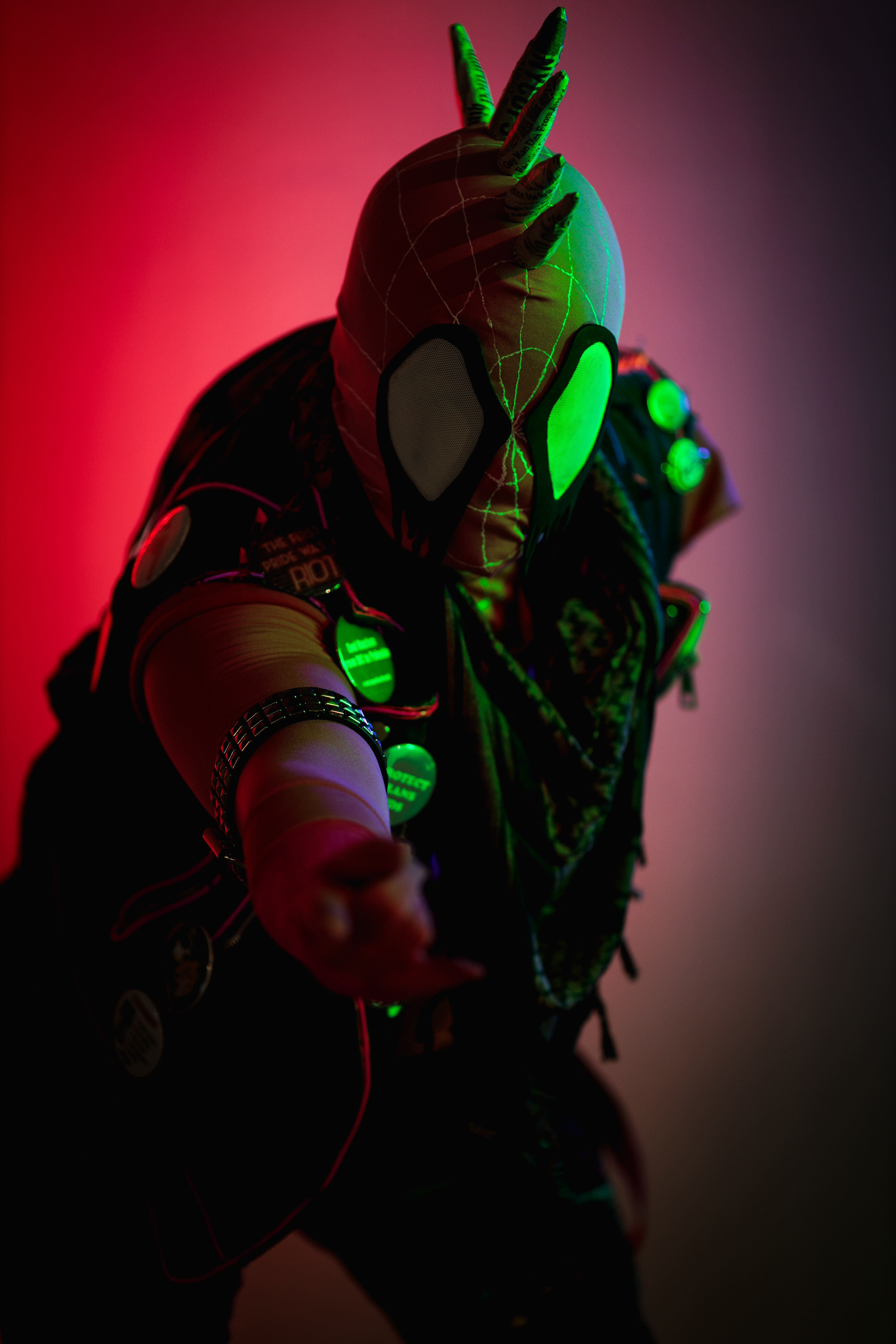 Spider-punk (pink palette) cosplayer wearing a pink keffiyeh illuminated by colorful light, pointing a "web-shooting" hand gesture at the camera