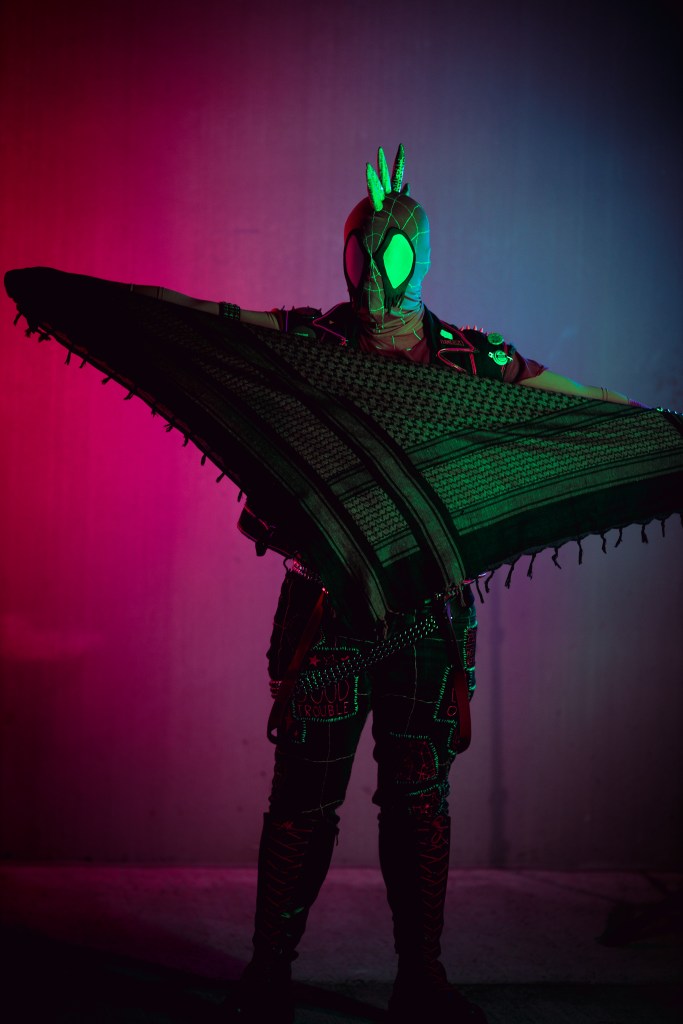 Spider-punk (pink palette) cosplayer holding a pink keffiyeh, illuminated by colorful light