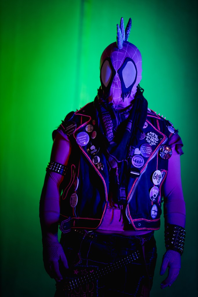 Spider-punk (pink palette) cosplayer wearing a pink keffiyeh, illuminated by colorful light
