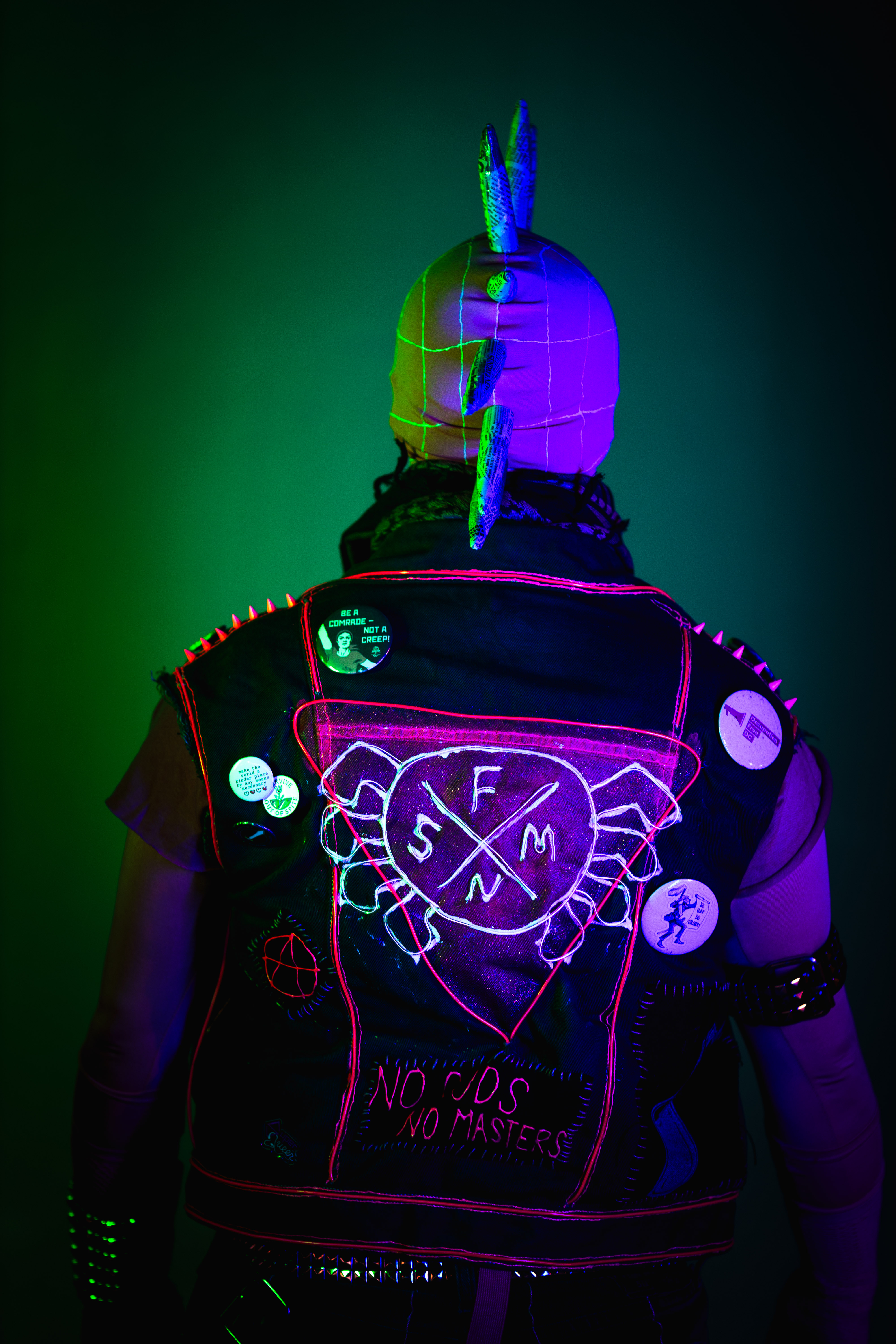 Spider-punk (pink palette) cosplayer wearing a pink keffiyeh illuminated by green light on the left and blue light on the right. Their back is turned, showing the "No Gods, No Masters" patch at the bottom of their vest, and the Spider-Punk logo superimposed over a glittery pink triangle.