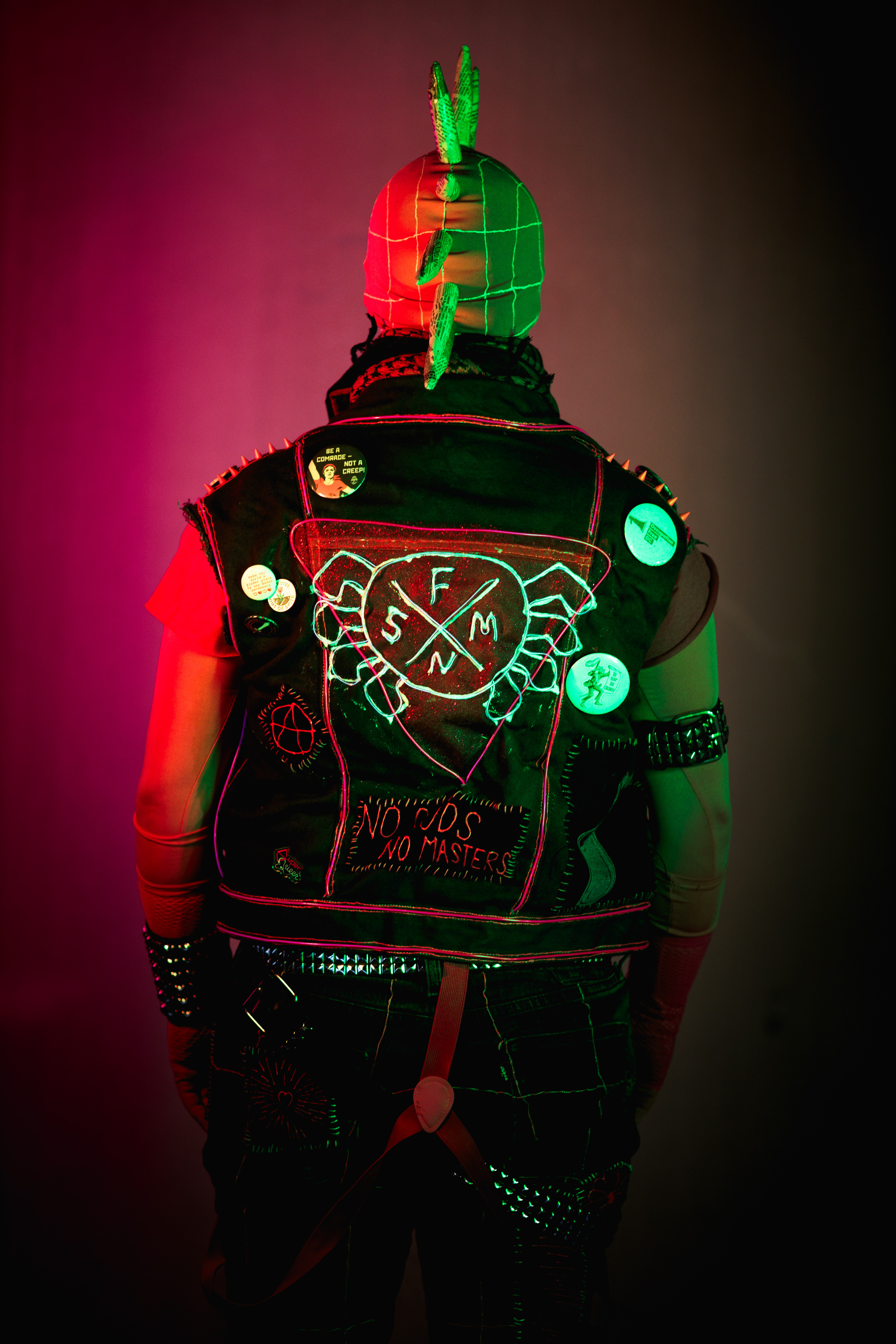 Spider-punk (pink palette) cosplayer wearing a pink keffiyeh illuminated by pink light on the left and warm light on the right. Their back is turned, showing the "No Gods, No Masters" patch at the bottom of their vest, and the Spider-Punk logo superimposed over a glittery pink triangle.