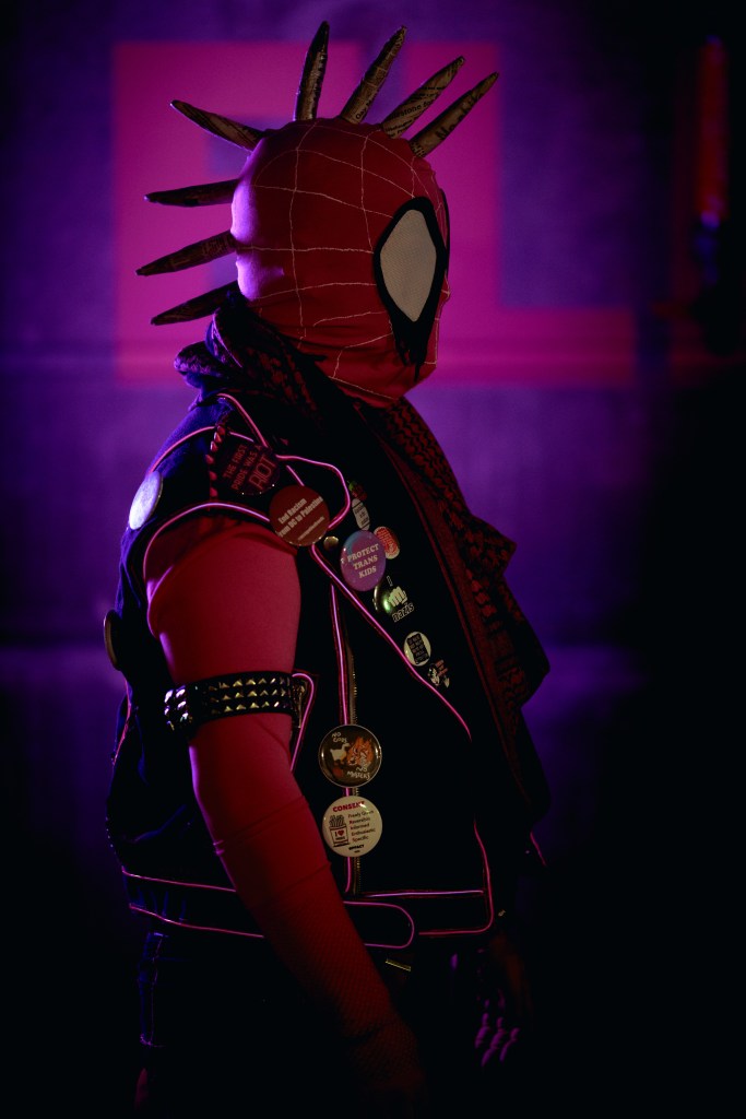 Spider-punk (pink palette) cosplayer wearing a pink keffiyeh, illuminated by colorful light, facing right