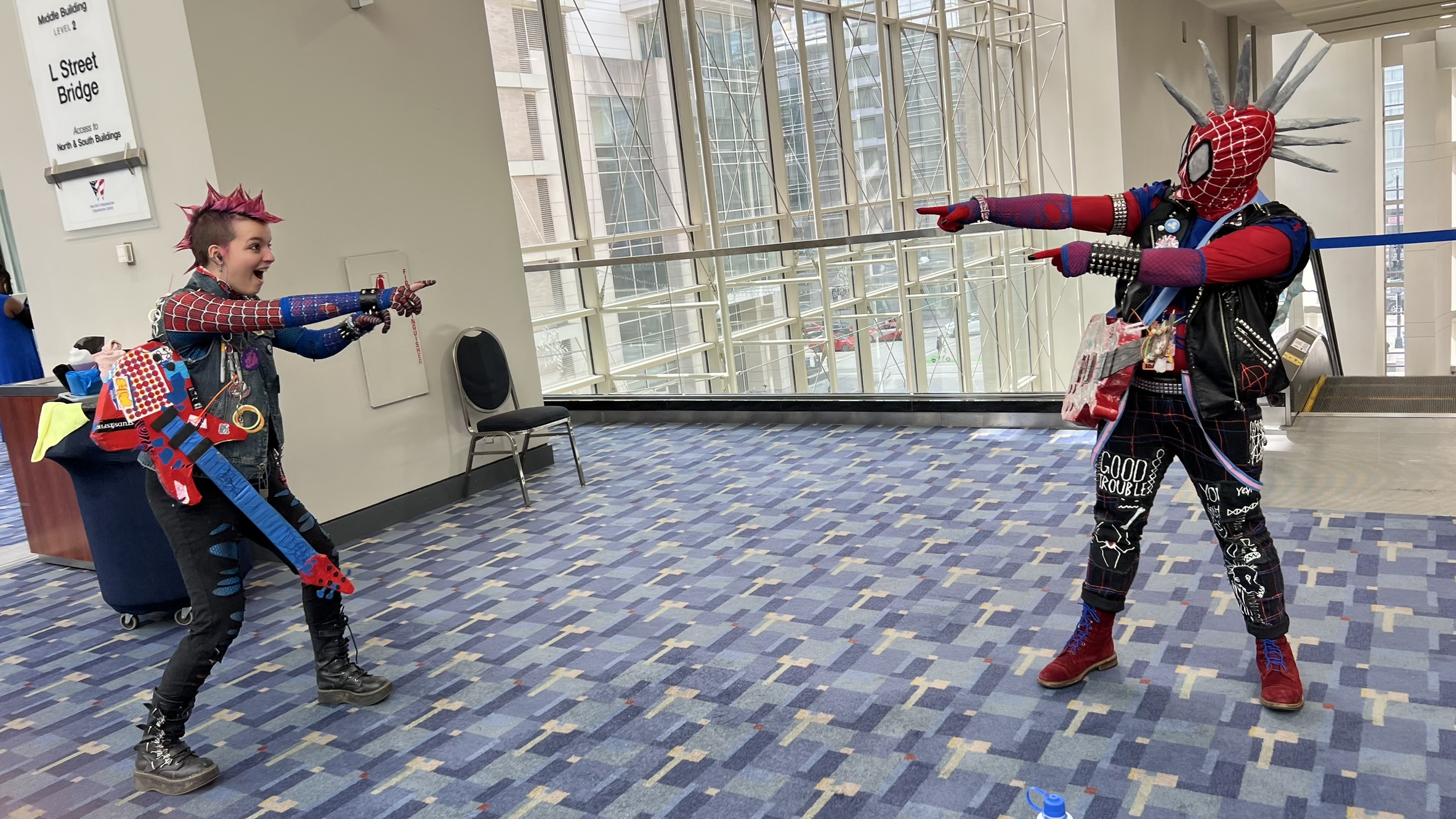 Two Spider-Punk cosplayers pointing at each other. One is masked, the other is unmasked and has a pink spiked mohawk.