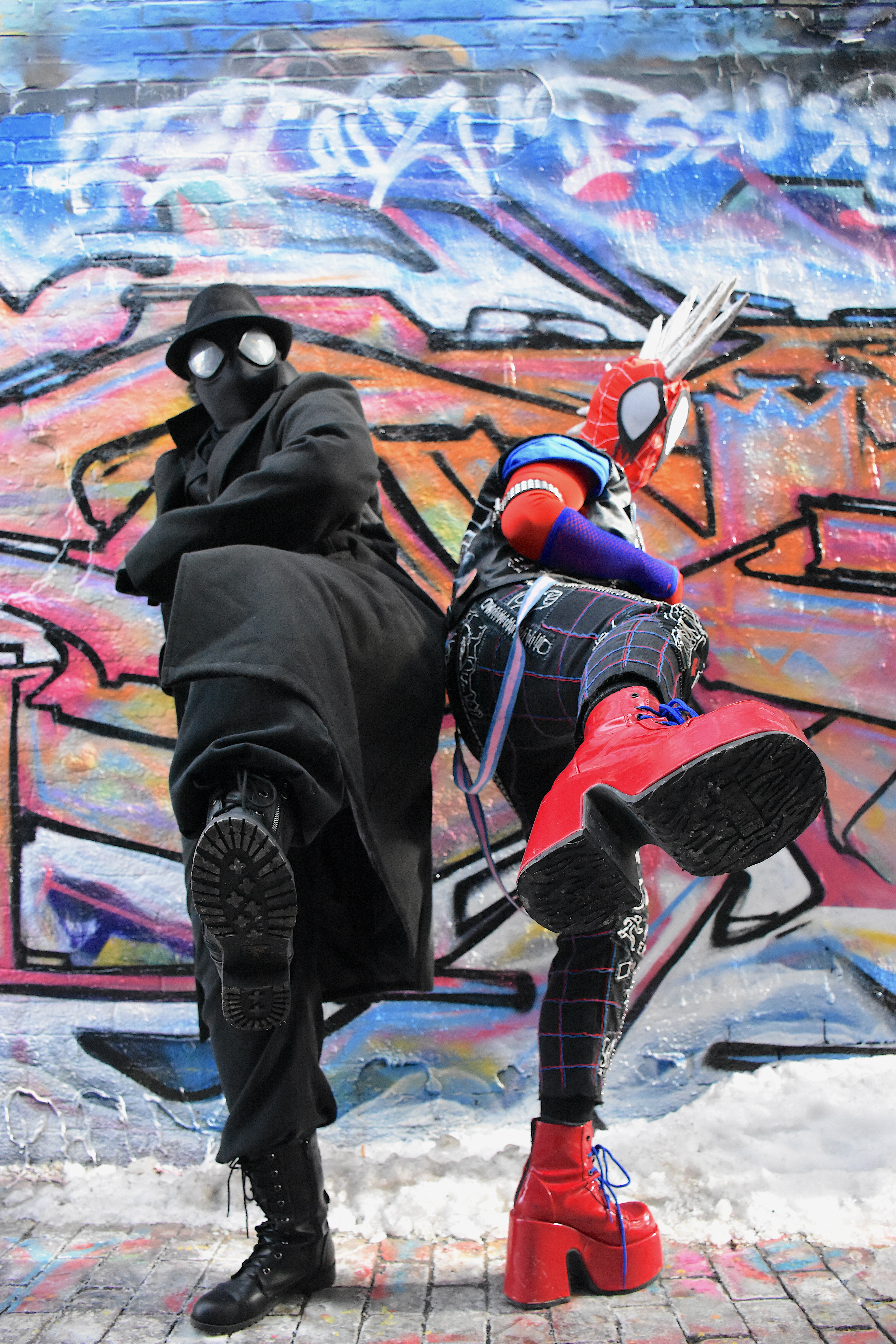 Spider-Man Noir and Spider-Punk cosplayers pose back-to-back in front of a graffiti mural, photographed mid-kick. There is snow on the ground.