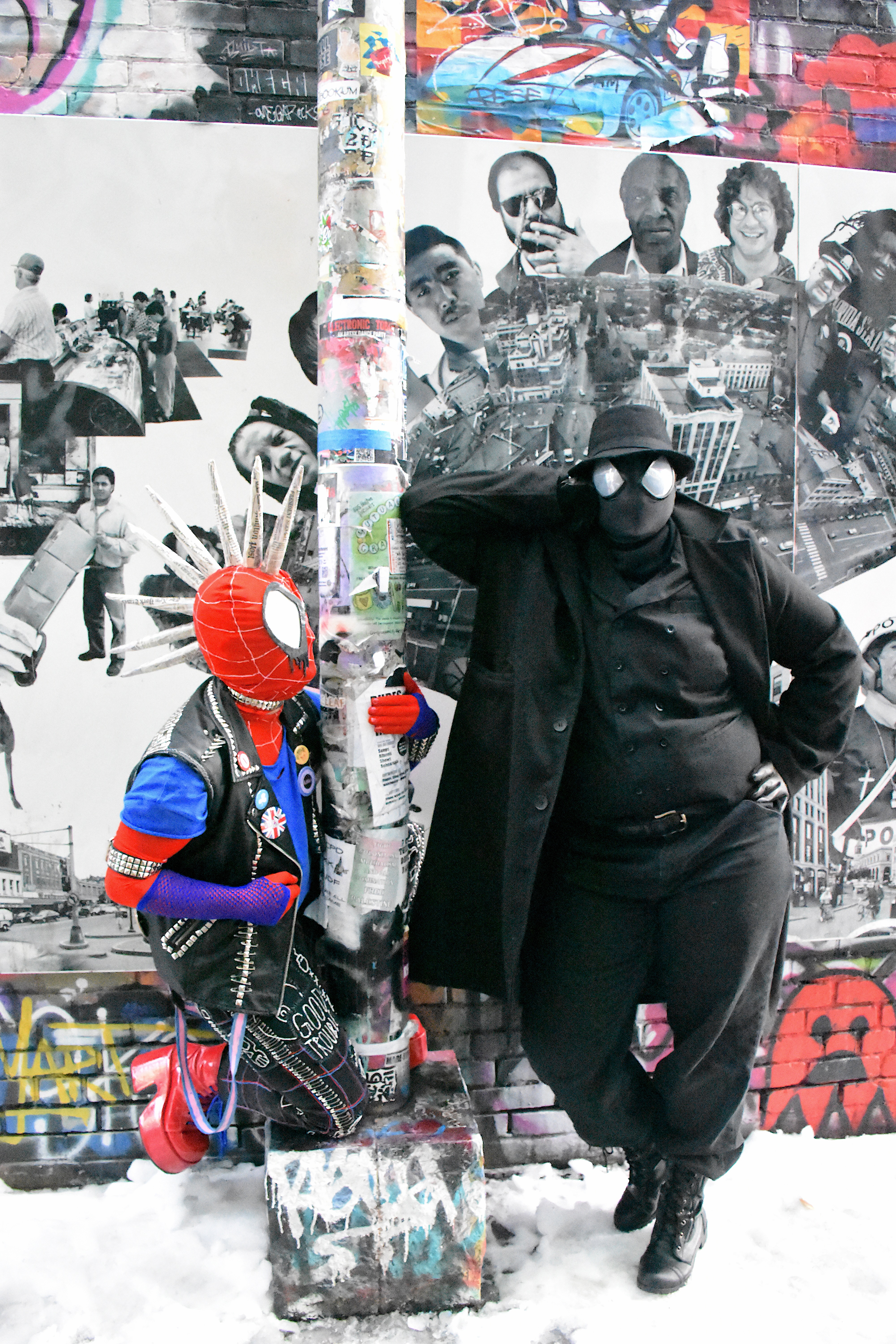 Spider-Man Noir cosplayer leans against a lamppost, looking at the camera. Spider-Punk cosplayer is perched on the lamppost base looking up at him. The lamppost is covered in flyers and stickers